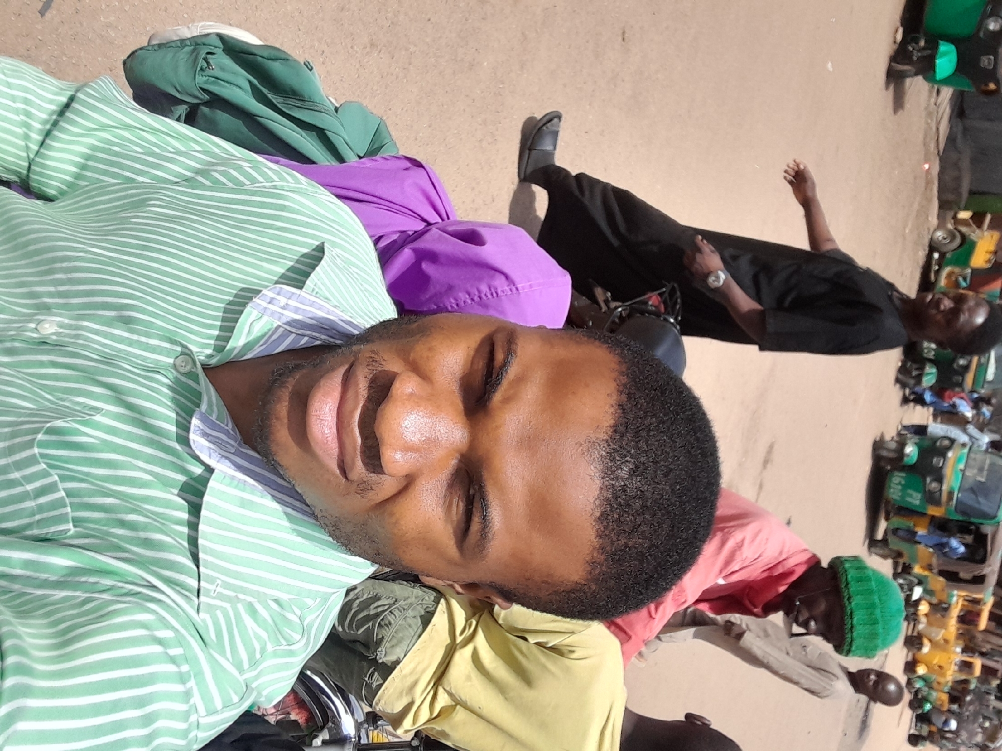 A medium close-up shot of a man wearing a light green and white striped button-down shirt, posing for the camera with his eyes closed. Behind him are various bags and clothing items including a green jacket, a purple bag, and a yellow shirt. A man in a black shirt is visible in the midground, his arm raised. In the background, there are several green and yellow motorized tricycles (Keke Napep) commonly used for transportation. A man with a green beanie is also visible amongst them. The scene is on a paved surface, suggestive of a street. Given the presence of Keke Napeps and the overall style, it is highly probable that this image was taken in Jos, Nigeria, where these vehicles are a common mode of transport.
