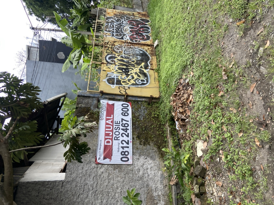 The image shows a gray concrete wall with a red and white sign attached to it. The sign advertises the sale of a property by Discovery Property with the contact number 0812 2467 600. The sign also reads "Dijual Rosie" which translates to "For Sale Rosie". The wall is adjacent to a rusty yellow metal gate with graffiti on it. The ground is covered in green grass and brown leaves. The scene is located in Lembang, Indonesia, at the coordinates -6.834 latitude and 107.603 longitude.