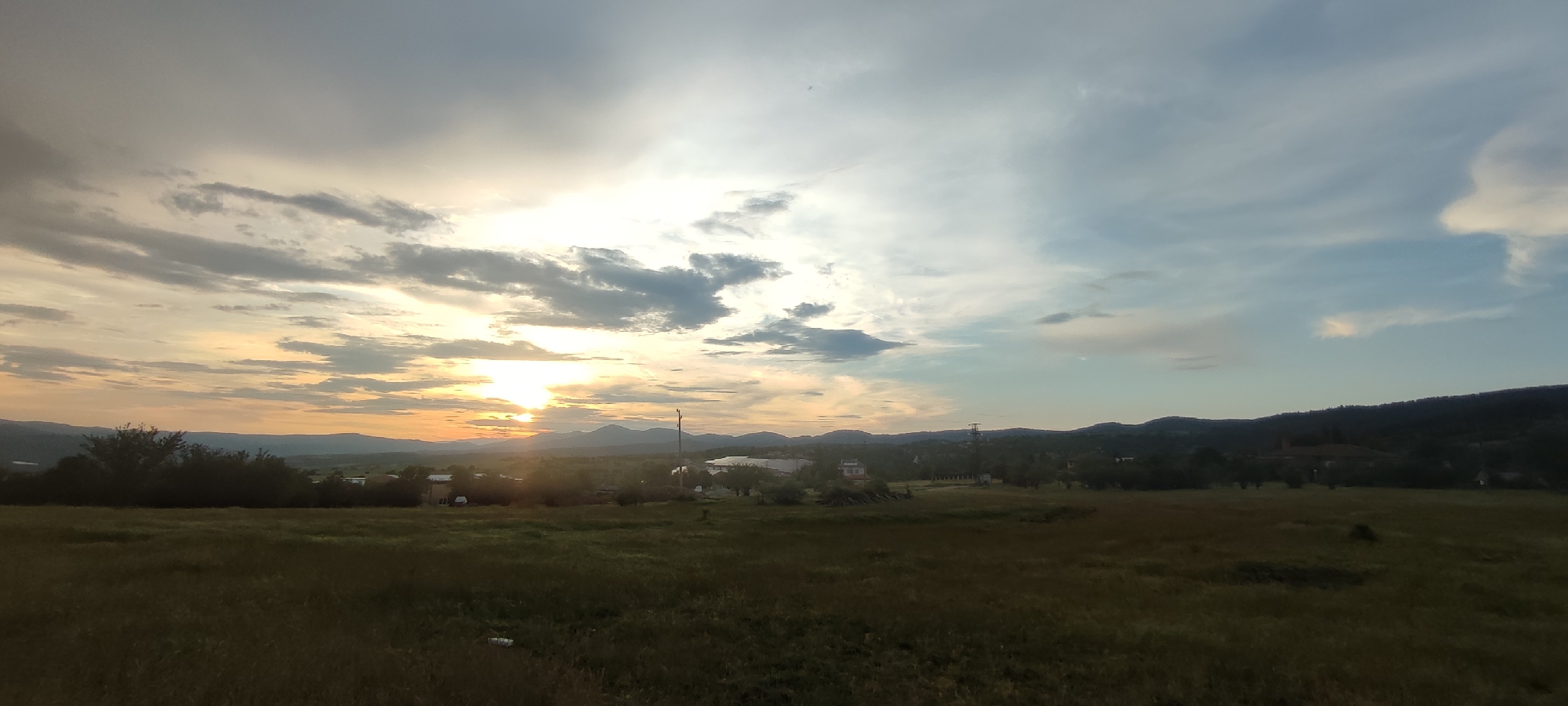 The image depicts a wide-angle landscape view of Momin Prohod, Bulgaria, at sunset. The foreground consists of a gently sloping field covered in dry, golden-brown grass, suggesting a rural or semi-rural setting. A few scattered white objects, possibly debris or rocks, are visible in the grass.
In the middle ground, a line of dark green trees and shrubs forms a dense barrier, obscuring a portion of the view beyond. Beyond the trees, hints of buildings and structures can be discerned, indicating the presence of a village or small town. A tall, slender tower, possibly a communication or observation tower, stands prominently on the right side of the middle ground. Further in the distance, rolling hills and mountains create a layered backdrop that recedes into the horizon.
The sky dominates the upper half of the image. The sun, a bright orb radiating warm yellow and orange light, is positioned near the center-left, partially obscured by soft, textured clouds. The clouds exhibit a range of colors from bright white and pale yellow near the sun to shades of gray and soft blue towards the edges and upper portion of the sky. The light from the setting sun casts a warm glow over the landscape, illuminating the clouds and creating a serene, atmospheric effect. The overall impression is one of a peaceful evening in a rural Bulgarian landscape. There are no people or discernible human activity in the image.
