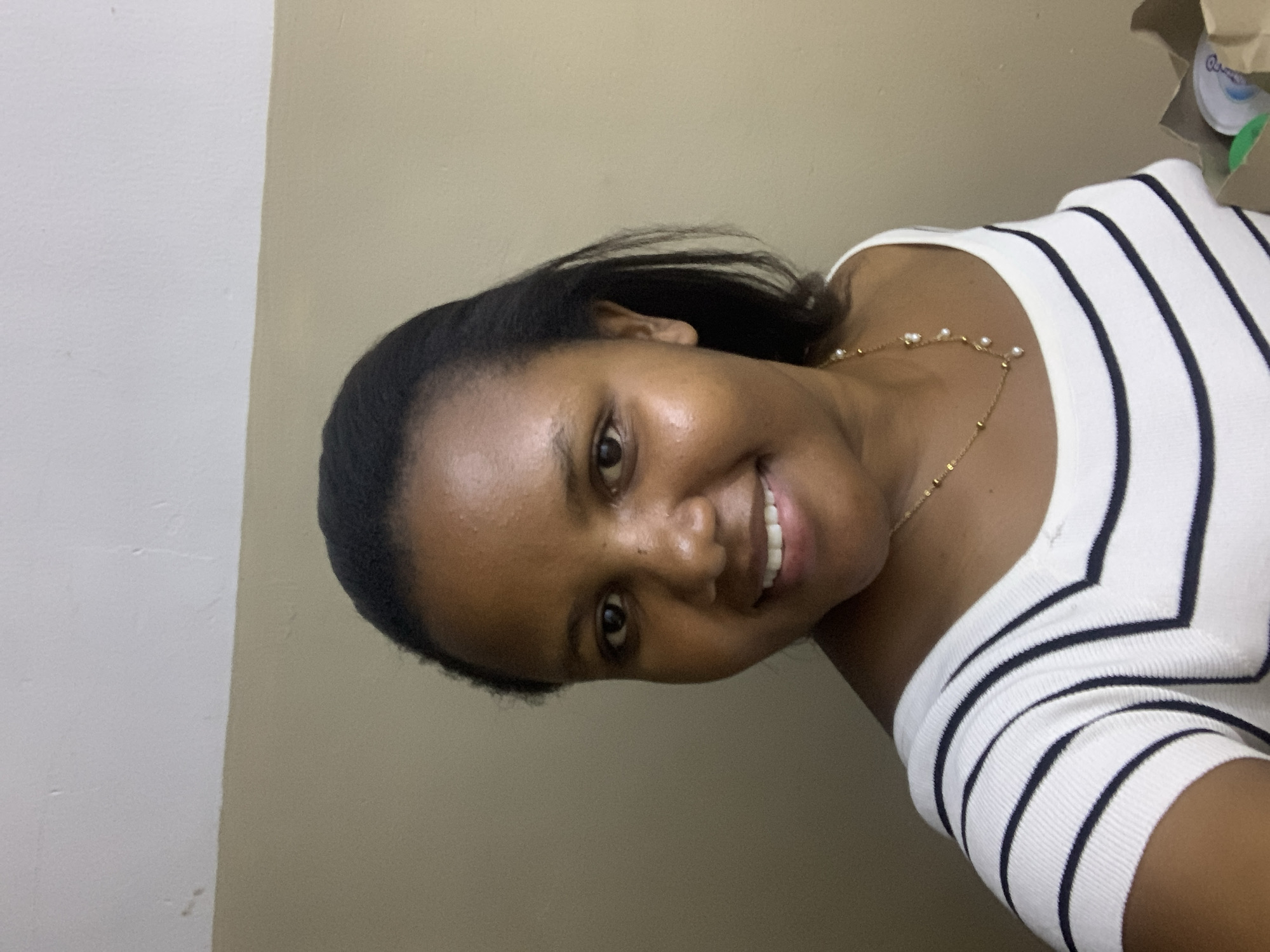 A woman smiles at the camera, wearing a white and black striped top and a gold necklace with pearls. She is positioned against a two-toned wall with the right side being beige and the left side being white. The image is an indoor shot with a slight angle. There's a paper bag with visible items at the upper right corner of the frame. It is impossible to ascertain the location in the city of Dar es-Salaam, Tanzania from the provided image.
