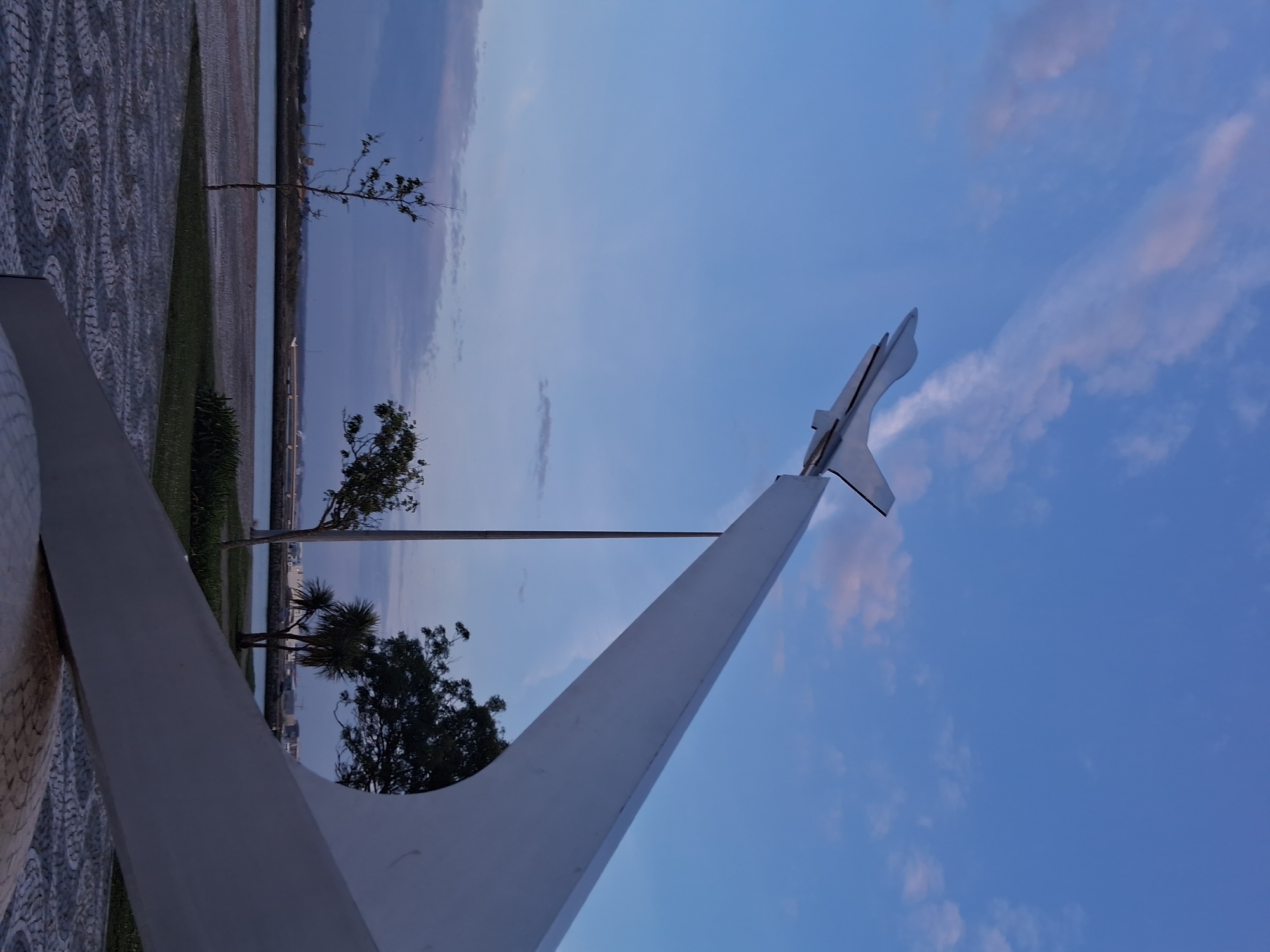 The image depicts the bronze marker "Monumento ao Aviador Gago Coutinho" located in Figueira da Foz, Portugal. The sky is blue with scattered clouds. The marker consists of a large white, triangular structure with a stylized airplane element at its peak. The structure appears to be made of concrete or a similar material. A thin metal pole extends vertically towards the top of the sky, presumably a part of the design. A paved walkway with a patterned surface is visible in the foreground, with a narrow strip of green grass along the edge. Behind the marker, a beach is seen, with water and a distant shoreline under a hazy sky. Several trees are visible in front of the beach, adding to the background scenery.