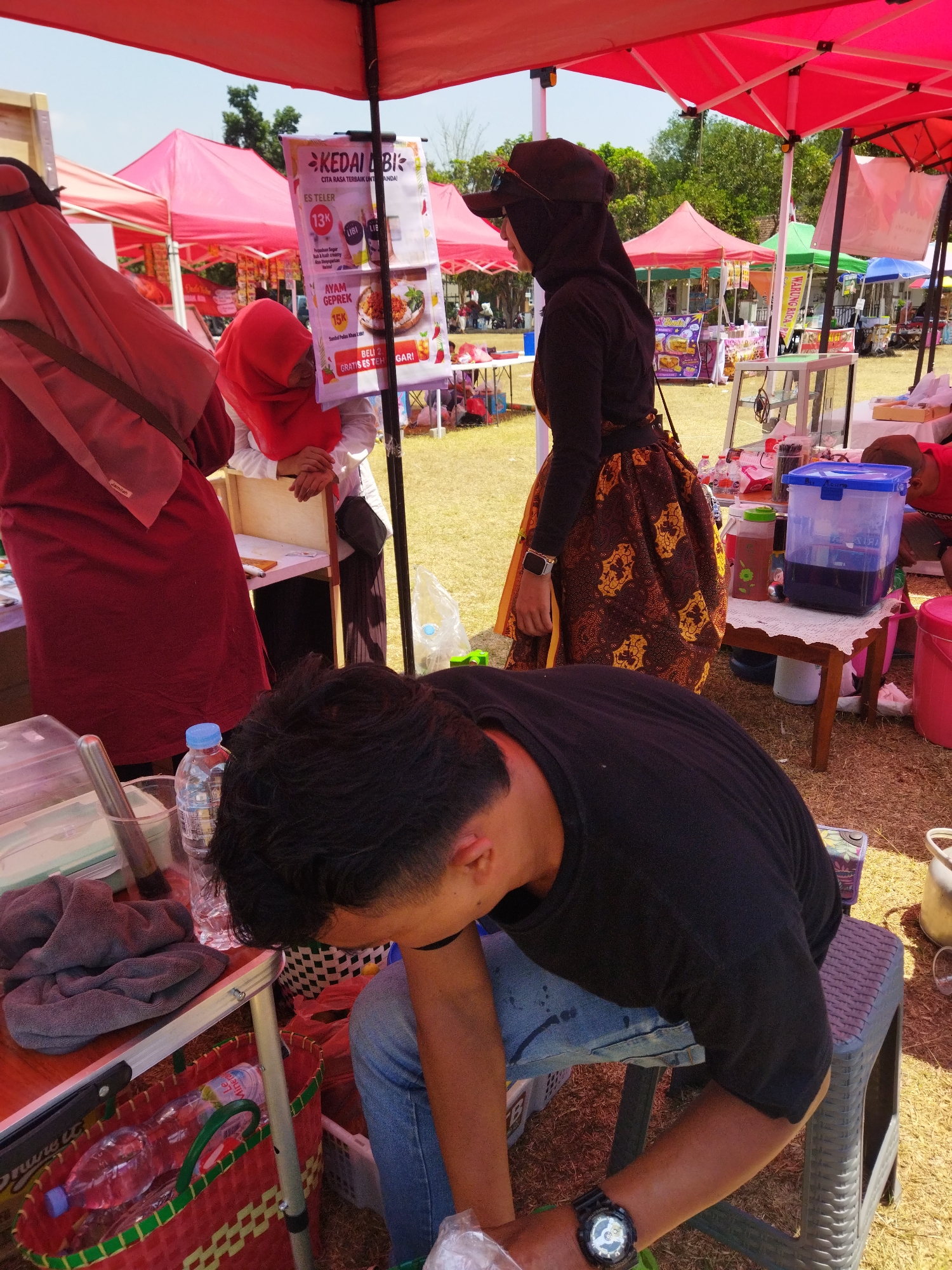 In Semarang, Indonesia, an outdoor market scene unfolds under a bright sky with scattered clouds. Red and pink tents offer shade, and the ground is covered in dry grass. In the foreground, a person wearing a black t-shirt and blue jeans leans over a table, resting their arm on their knee. A black watch is visible on their wrist. To their left, a red woven basket holds plastic bottles of water. Further back, a person in a dark headscarf and patterned skirt stands near a display of food items. To the left, another individual in a red headscarf and a maroon dress is partially visible. Behind them, various stalls with food and drink offerings are set up, suggesting a lively marketplace atmosphere.