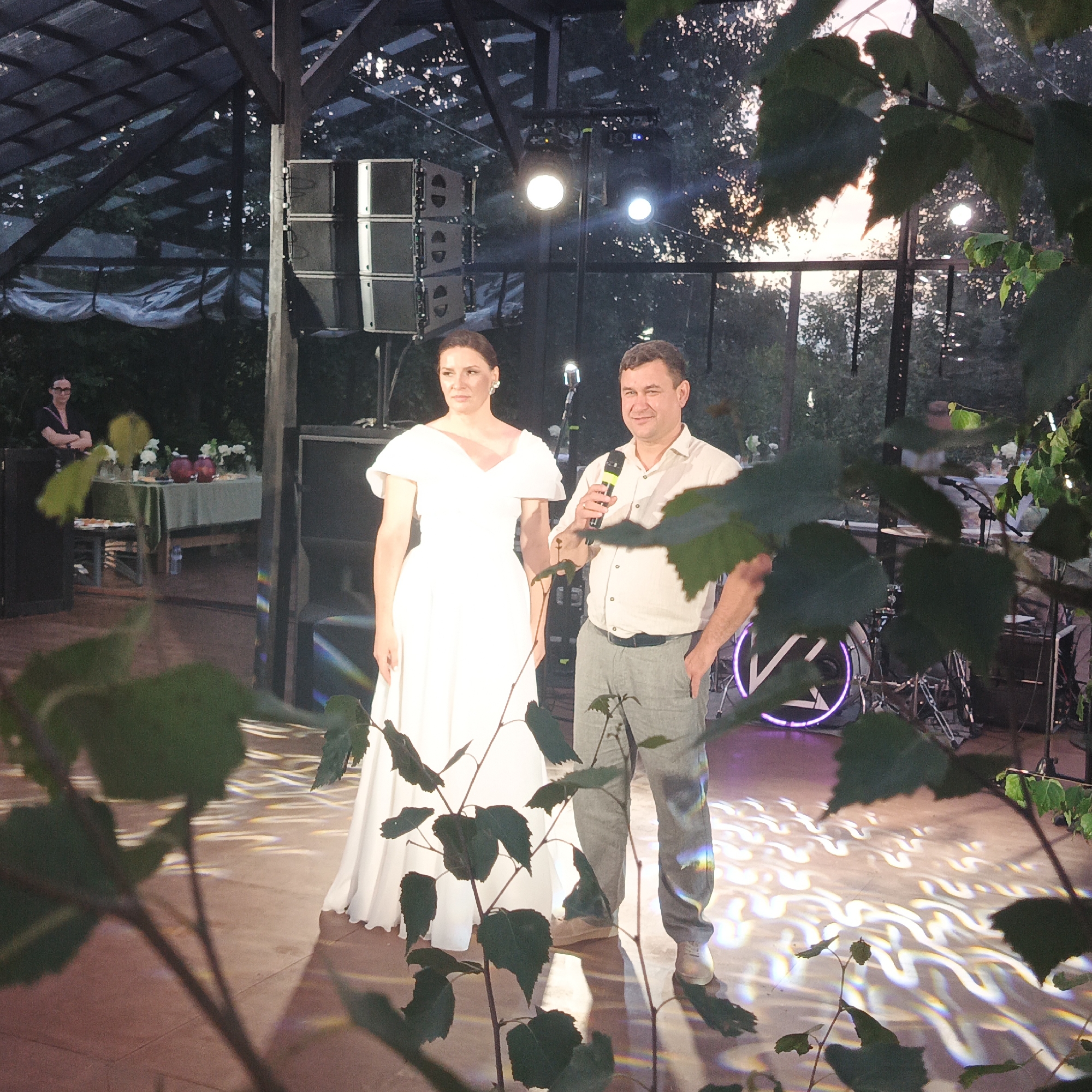 The image depicts an outdoor event or venue in Svetloe, Russia, possibly a wedding or concert, with elements suggesting an evening setting. In the foreground, the lower portion of several plants with green leaves obscures the view, creating a natural framing effect.
The central focus is on a man and a woman standing on a stage or performance area. The woman, positioned on the left, is wearing a long, white, formal gown with puffy sleeves and a V-neckline. She has her hair pulled back and is wearing earrings. Her expression is neutral, and she is looking slightly off to the side. The man, to her right, is holding a microphone and speaking. He is dressed in a light-colored collared shirt, grey trousers, and light-colored shoes. He appears to be gesturing with his left hand, which is tucked into his trouser pocket, and his right hand is holding the microphone. He has a slight smile on his face.
Behind the couple, a stack of large speakers is visible on the left, indicating a sound system. Lights are illuminating the stage area, casting distinct wavy patterns on the floor, some of which are iridescent, suggesting a prismatic effect. In the background, there is a transparent roof structure with dark beams, and through it, the sky appears to be transitioning from daylight to twilight, with dappled light filtering through trees. Further in the background, a table laden with floral arrangements and decorative items is set up, suggesting a catered event. To the far left, a person with dark hair and glasses is standing behind a railing, observing the scene. A drum set with a circular illuminated logo is visible to the right of the man, implying a musical performance. The overall atmosphere is festive, yet intimate, with natural elements intertwined with a stage setup.