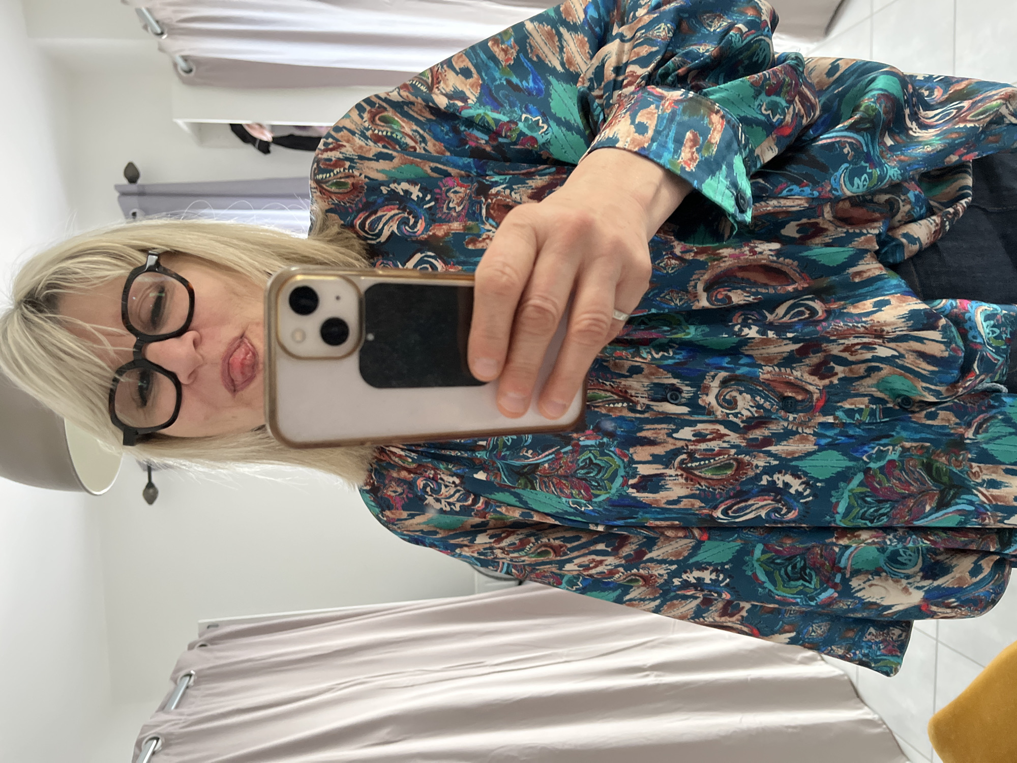An indoor mirror selfie taken in Chambœuf, France, depicts an individual from the chest up. The person, with blonde hair and wearing dark, round-rimmed glasses, is shown sticking their tongue out while holding a light-colored smartphone with a dual-camera system. They are wearing a loose-fitting, long-sleeved blouse featuring a multi-colored abstract and paisley pattern in shades of teal, blue, brown, red, and beige, along with dark lower garments. A silver-toned ring is visible on the ring finger of the hand holding the phone. The background, reflected in the mirror, consists of a white wall, a grey bowl-shaped ceiling light fixture, and light-beige curtains on metal rods on both sides. A white tiled floor is visible at the bottom, alongside a partial view of a yellow upholstered object in the lower left and a white shelving unit with dark clothing on the right.