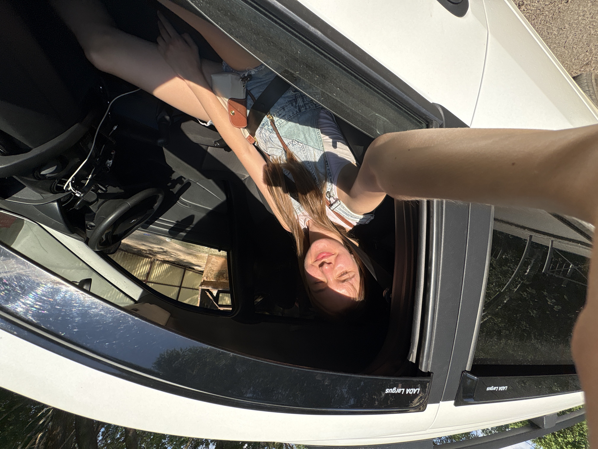 A young woman is taking a selfie from inside a white Lada Largus car. The car is in Perm, Russia, based on the Cyrillic inscription on the window trim. The woman is wearing denim overalls and has a small purse strapped across her chest. The car interior is black. Exterior details, including a partially visible building and trees, suggest a sunny outdoor setting.