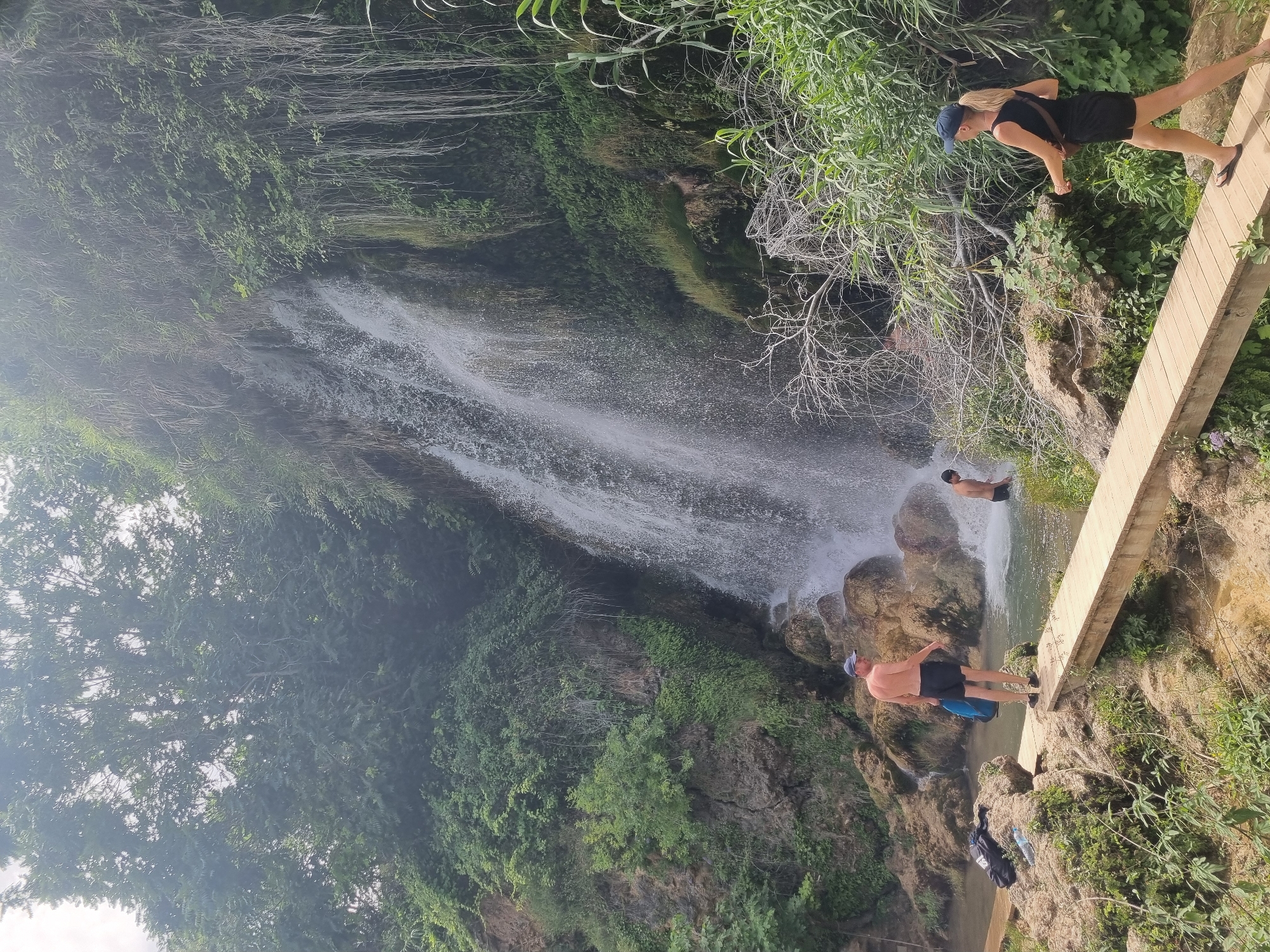 A group of people are at a waterfall in the city of Anna, Spain. In the foreground, a woman with blonde hair, wearing a blue cap, black tank top, and black shorts, walks on a wooden bridge. To her left, a man with dark hair, wearing a blue cap and black shorts, stands on a rock near the waterfall. Further left, a man with dark hair and a blue cap, wearing black shorts, stands on a wooden walkway. In the background, a waterfall cascades down a rocky cliff face covered in greenery. The scene appears to be a natural park or recreational area.