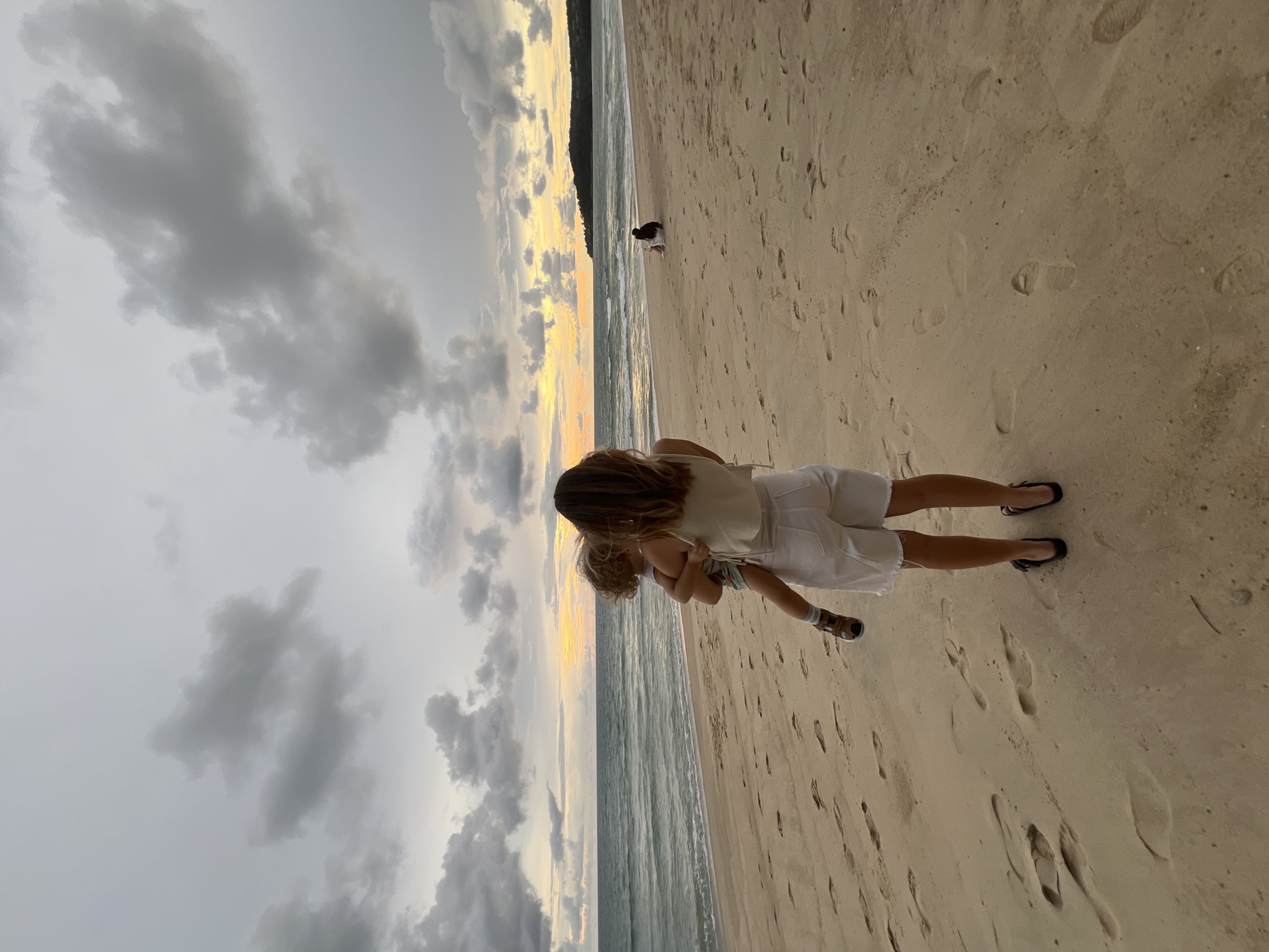 A woman holds a toddler on a sandy beach at dusk. The woman, facing away from the camera, wears a cream-colored tank top and white shorts. She has long brown hair. The toddler, also facing away, has blond curly hair and wears a patterned outfit. The beach is covered in footprints. The ocean is to the left, with gentle waves. The sky is dramatic, with dark gray clouds and a hint of orange and yellow sunset. In the distance, another person sits on the sand, and a dark, tree-covered hill is visible along the coastline.