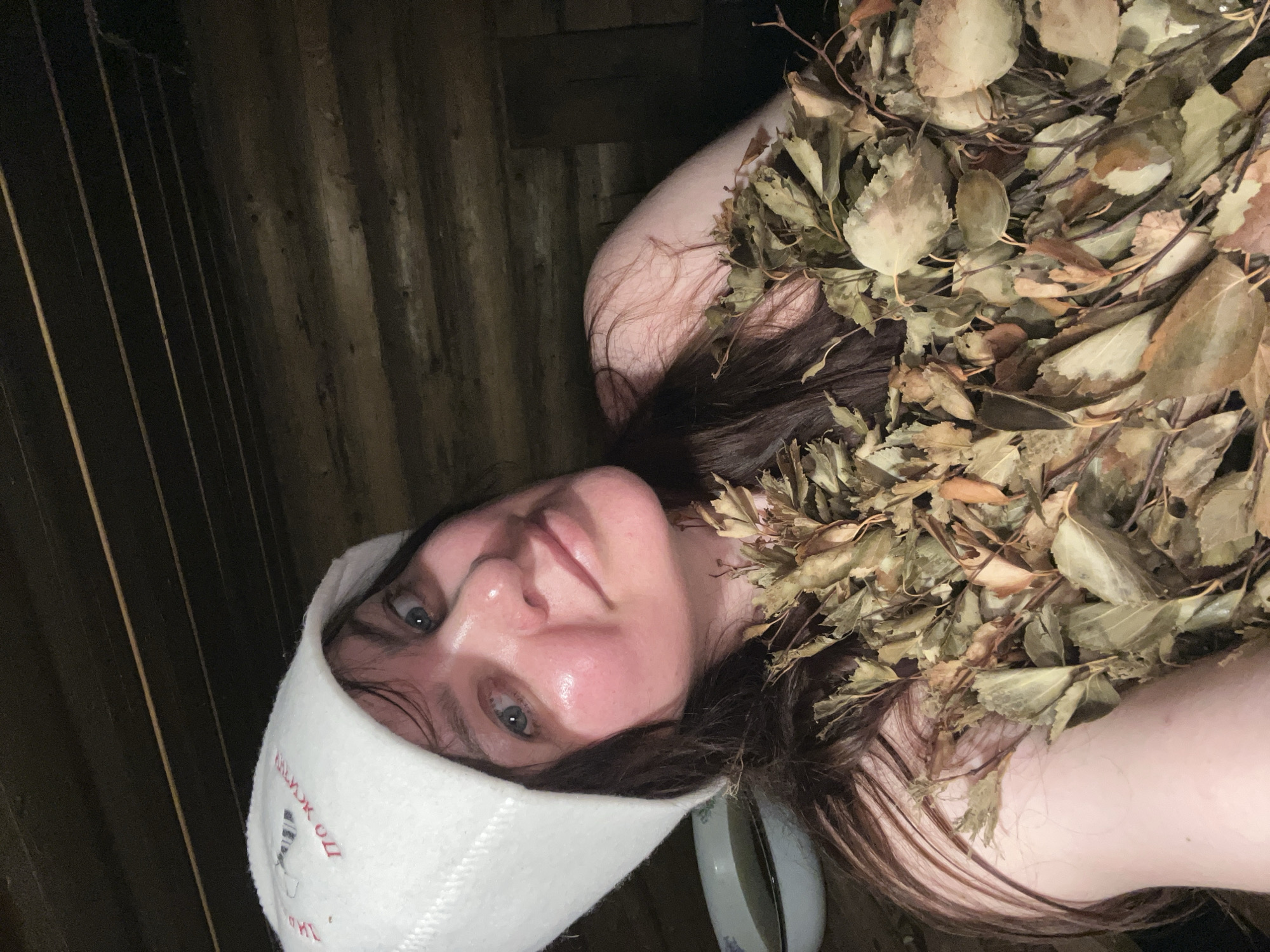 A woman is lying down, wearing a white sauna hat with text and a logo, and covered in dried leaves. The background appears to be wood paneling, possibly inside a sauna. A round object resembling a bowl is partially visible near her head. The location is purported to be in Protasy, Russia, though this cannot be independently verified from the image alone.