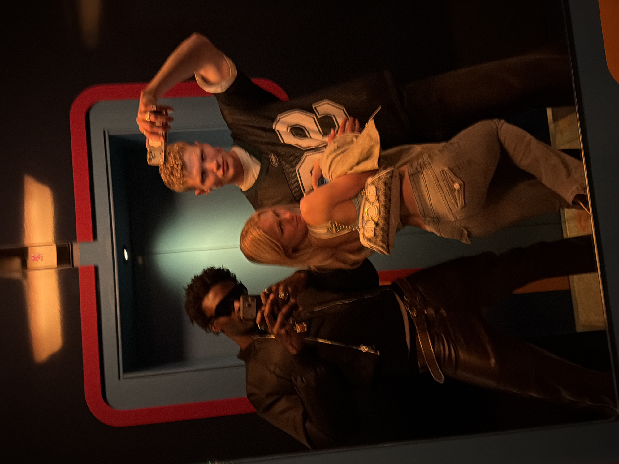 Three individuals are reflected in what appears to be a mirror or reflective surface encased in a frame with red trim. The frame is set against a blue or grey background, suggesting a modern or industrial setting. The reflection shows the top half of three people.
One individual, on the left, is a man with fair skin and light-colored hair, wearing a black sports jersey with the number 82. He is holding a small object, possibly a phone or wallet, above his head.
Another person, positioned between the first man and a third person, is a woman with blonde hair. She is wearing a light-colored top with what looks like metal adornments around the chest.
A third person, located on the right, is a man with dark skin wearing a black jacket, sunglasses, and several rings on his fingers. He is holding a smartphone up to take the picture of all three.
The angle and reflection suggest the photo is being taken in a public space or potentially within a display or installation.
It is impossible to determine with certainty the precise location in London, UK without additional information or visual cues.