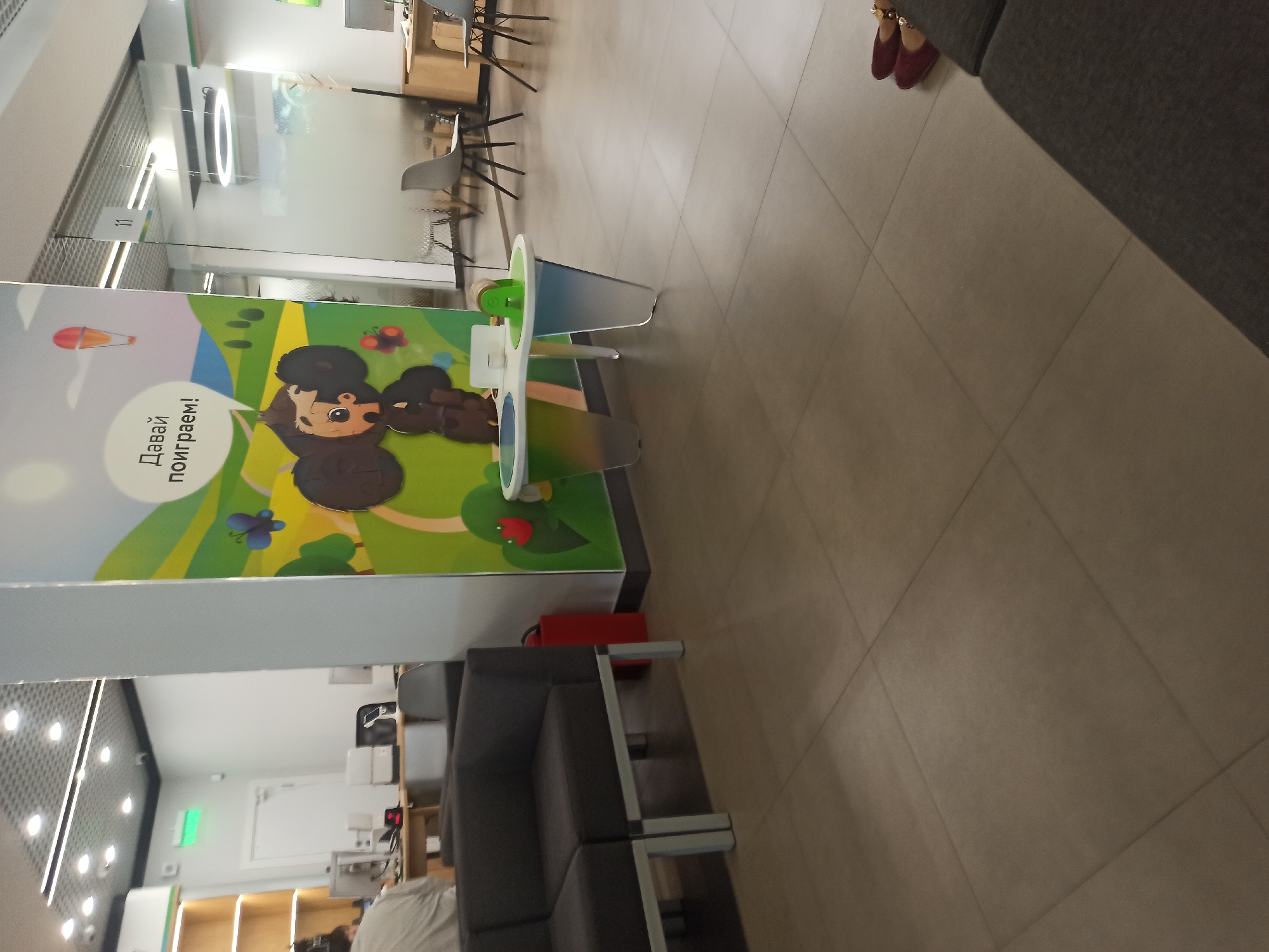 The image depicts the interior of a modern office space or waiting area, likely in Atabe, Georgia. The floor is tiled with large, light-brown square tiles. A dark grey sectional sofa is positioned in the foreground on the left. To the right of the sofa, a large, colorful mural featuring a cartoon character with large ears and a speech bubble with Russian text is prominent. Next to the mural, a white and gradient colored table with circular indentations and a green toy is situated. In the background, glass partitions and modern office furniture, including chairs and desks with computers, are visible. The lighting appears to be overhead fluorescent and circular recessed lighting. A red fire extinguisher is mounted on the wall near the sofa. The overall impression is of a clean, well-lit, and contemporary commercial environment. There are no visible people in the immediate foreground or engaged in specific actions, though shadows suggest movement or presence just outside the frame. The environment suggests a place designed for customer service or as a waiting area.