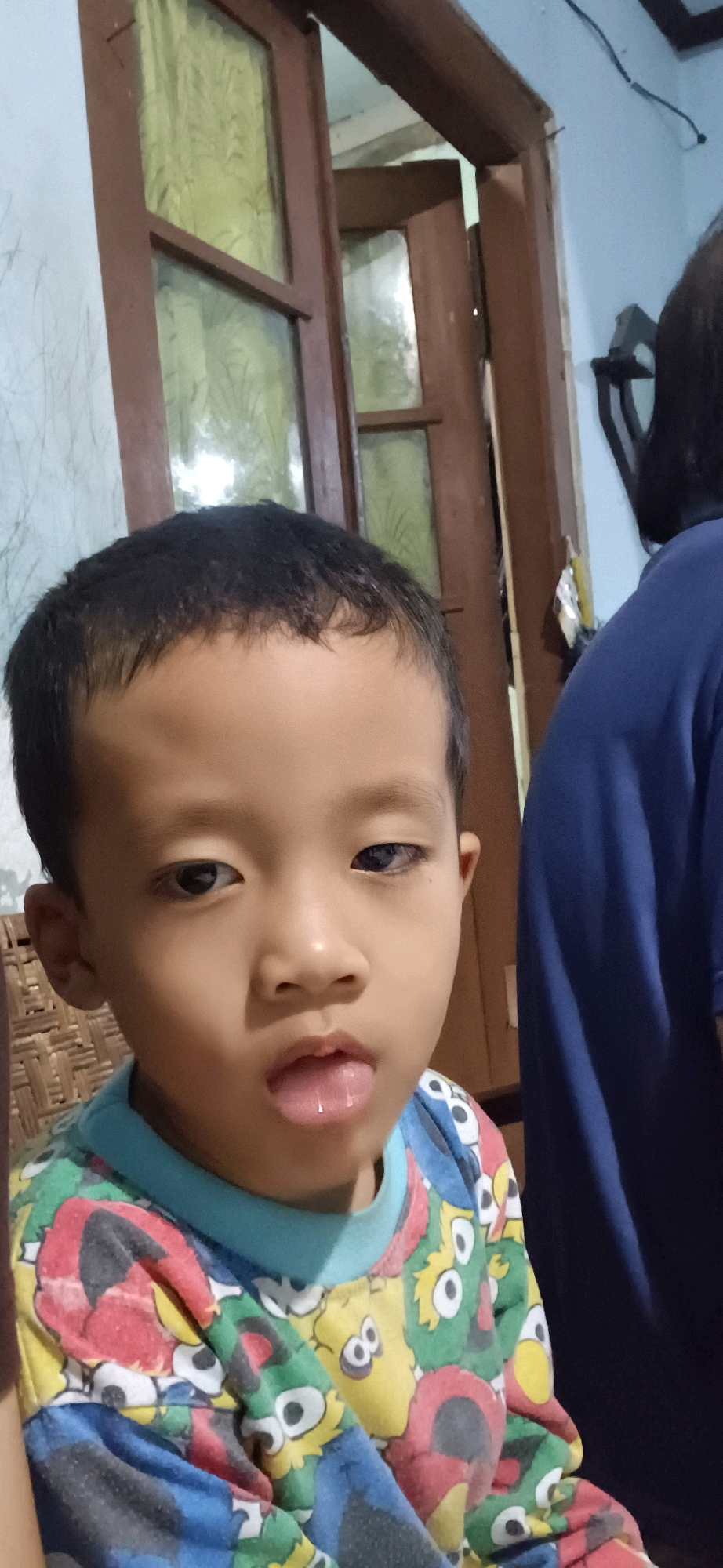 The image depicts a close-up, eye-level portrait of a young East Asian child, appearing to be between 4 and 6 years old. The child has short, dark hair and fair skin. Their mouth is slightly open, with the tongue extended and glistening. The child is wearing a long-sleeved t-shirt featuring a colorful pattern of cartoon characters, including Cookie Monster and Elmo from Sesame Street. The background is dimly lit and out of focus. To the left of the child, a woven texture, possibly from furniture, is visible. Behind the child, a wooden door frame with glass panes is present, suggesting an indoor setting. On the right side of the image, a portion of another person, wearing a dark blue garment, is visible. The lighting appears to be artificial and somewhat harsh, casting shadows on the child's face. The overall impression is of a candid snapshot taken indoors. While the prompt specifies a location in Wonokerso, Indonesia, there are no visible elements within the image itself that definitively confirm this geographical context. The scene could plausibly be in any residential setting with similar architectural features and interior decor.
