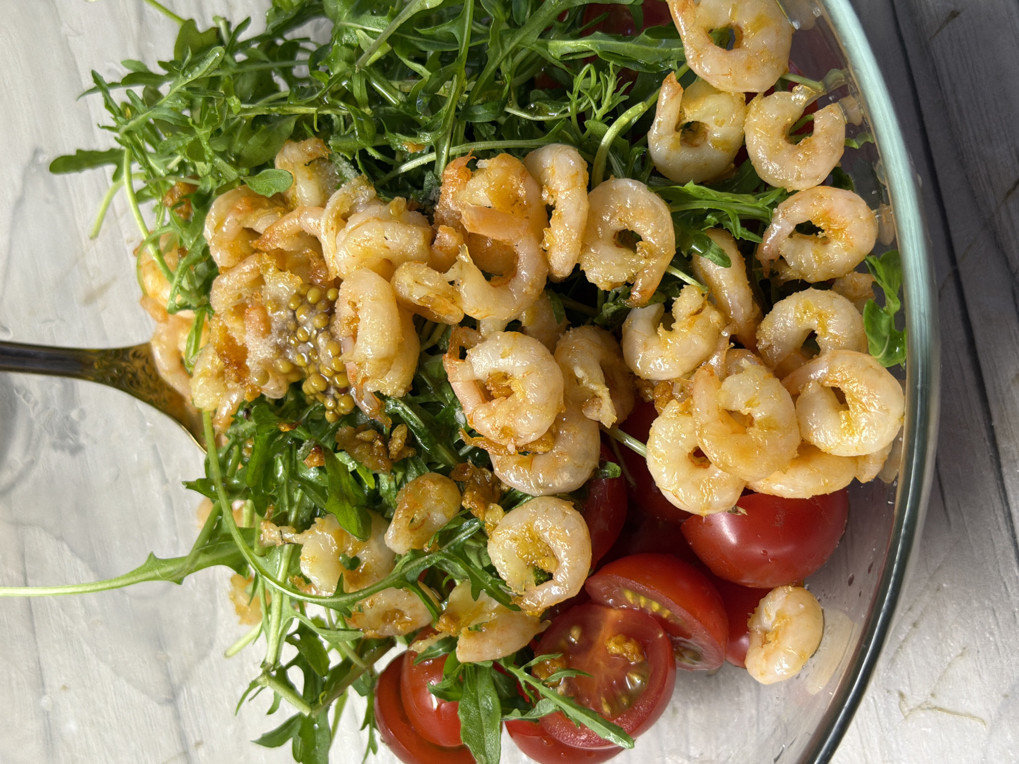 A close-up, overhead shot displays a glass bowl filled with a fresh salad composed of arugula, cherry tomatoes, and cooked shrimp. The arugula leaves are a vibrant green, some wilted, and mixed with diced, bright red cherry tomatoes. A generous portion of small, pinkish-orange shrimp are scattered throughout the salad, some appearing to be coated in a light dressing or seasoning. A silver fork is partially submerged in the salad, suggesting it's being served or prepared. The salad is presented on a light-colored, textured surface that resembles a wooden table or countertop. The lighting is bright and natural, highlighting the textures and colors of the food. There are no people visible in the image. The context of Chaikovsky, Russia, is not discernable from the visual information provided in the image.