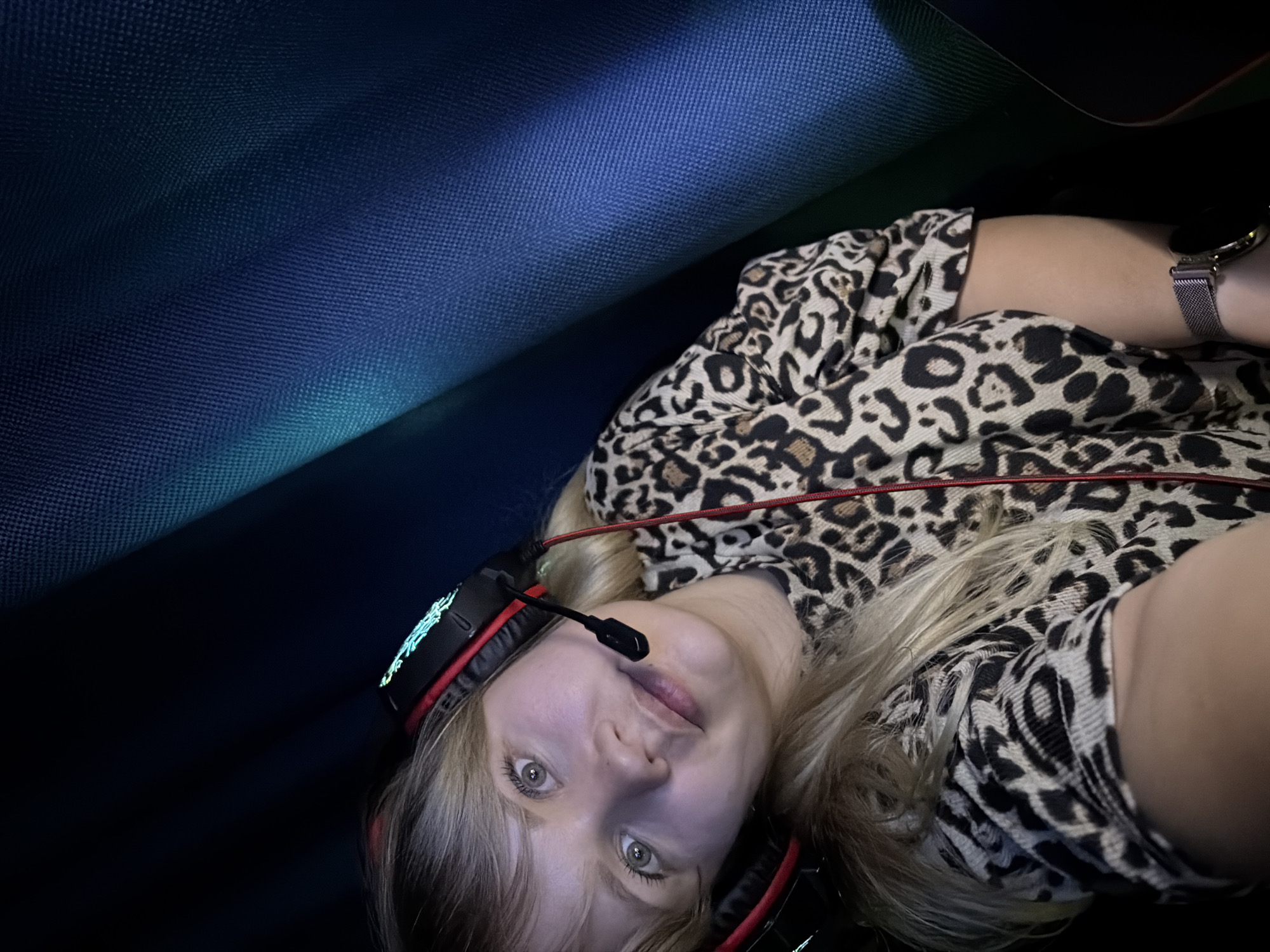 An upward-angled, close-up shot captures a young individual, likely in Chaikovsky, Russia, reclining indoors in a dimly lit setting. The subject, with blonde hair and light-colored eyes, looks directly at the camera with a neutral to slightly curious expression. They are wearing a black and brown leopard print top and a gaming headset with a microphone boom. The headset features glowing blue-green accents on the earcups or headband and has red cabling visible. A metallic mesh-band watch is visible on their left wrist.
The background consists of dark blue and black textured fabric, possibly part of a gaming chair, couch, or mat, suggesting an intimate, personal space. The ambient lighting is low, with some light sources illuminating the subject's face from below or the side, creating a soft glow on their skin. The overall atmosphere suggests an evening or nighttime setting, potentially during a gaming session or online interaction given the presence of the headset. No text is visible in the image.