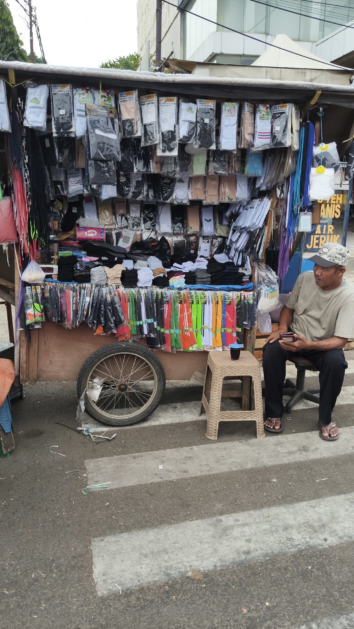 A man wearing a camouflage baseball cap, a light-colored t-shirt, black pants, and brown flip-flops sits on a chair in front of a street stall. He is holding a smartphone in his right hand and looking to his left. The stall, which is likely located in South Jakarta, Indonesia, is laden with various types of socks. The socks are displayed in clear plastic packaging and hung from racks. There are also multi-colored strings of fabric or ribbons hanging from the stall's canopy. A bicycle wheel is visible in the foreground, to the left of the man. A small beige plastic stool sits in front of the stall, with a small cup on it. The stall is set up on a street with a pedestrian crossing marked by white stripes. The background shows a building with a white facade and a light-colored roof.
