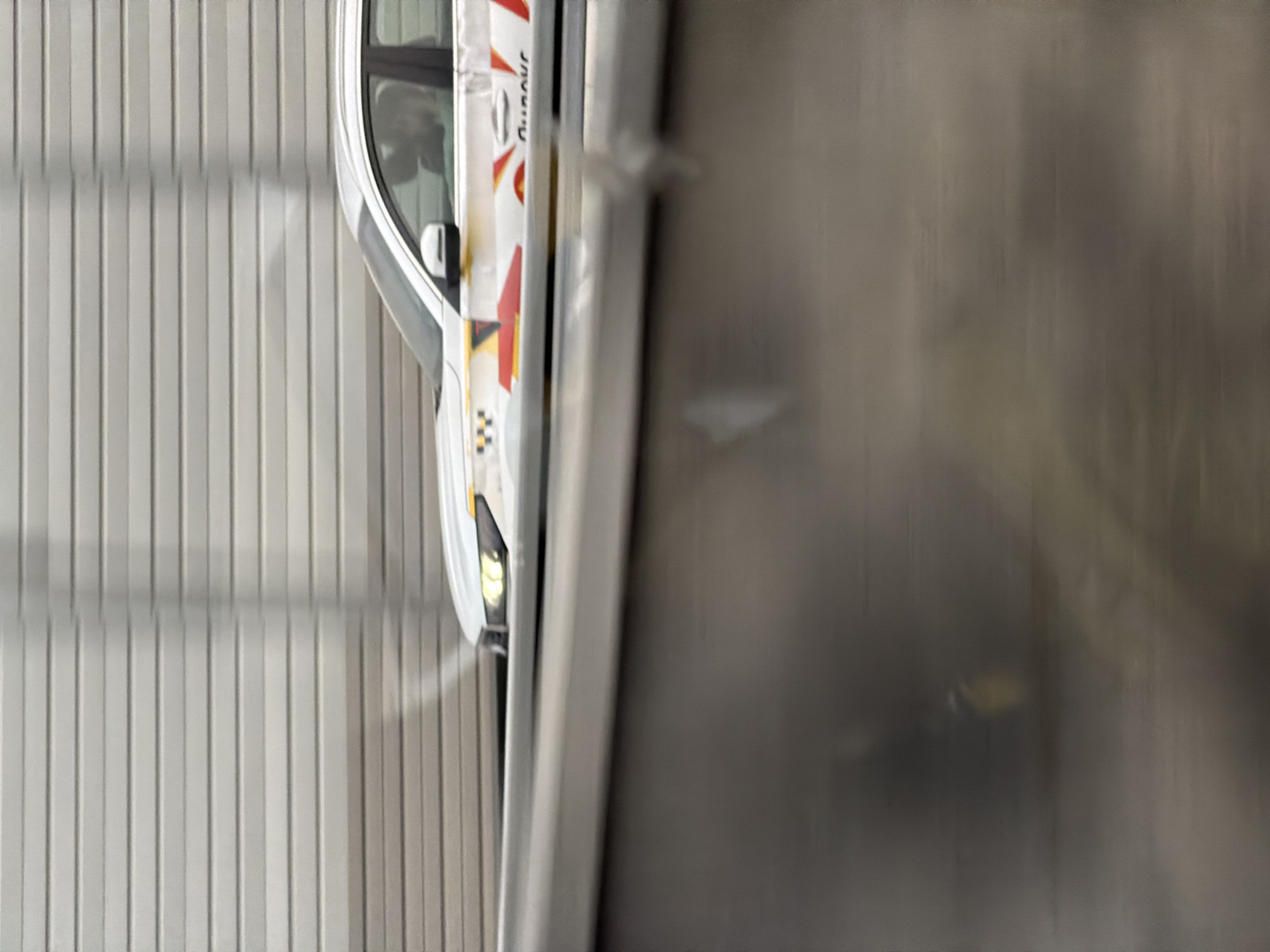Here is a forensic description of the image:
The image shows the side of a white taxi parked beside a wall. The taxi has orange and red decals on its side. The wall appears to be made of metal panels. The location of the scene is not evident from the image.