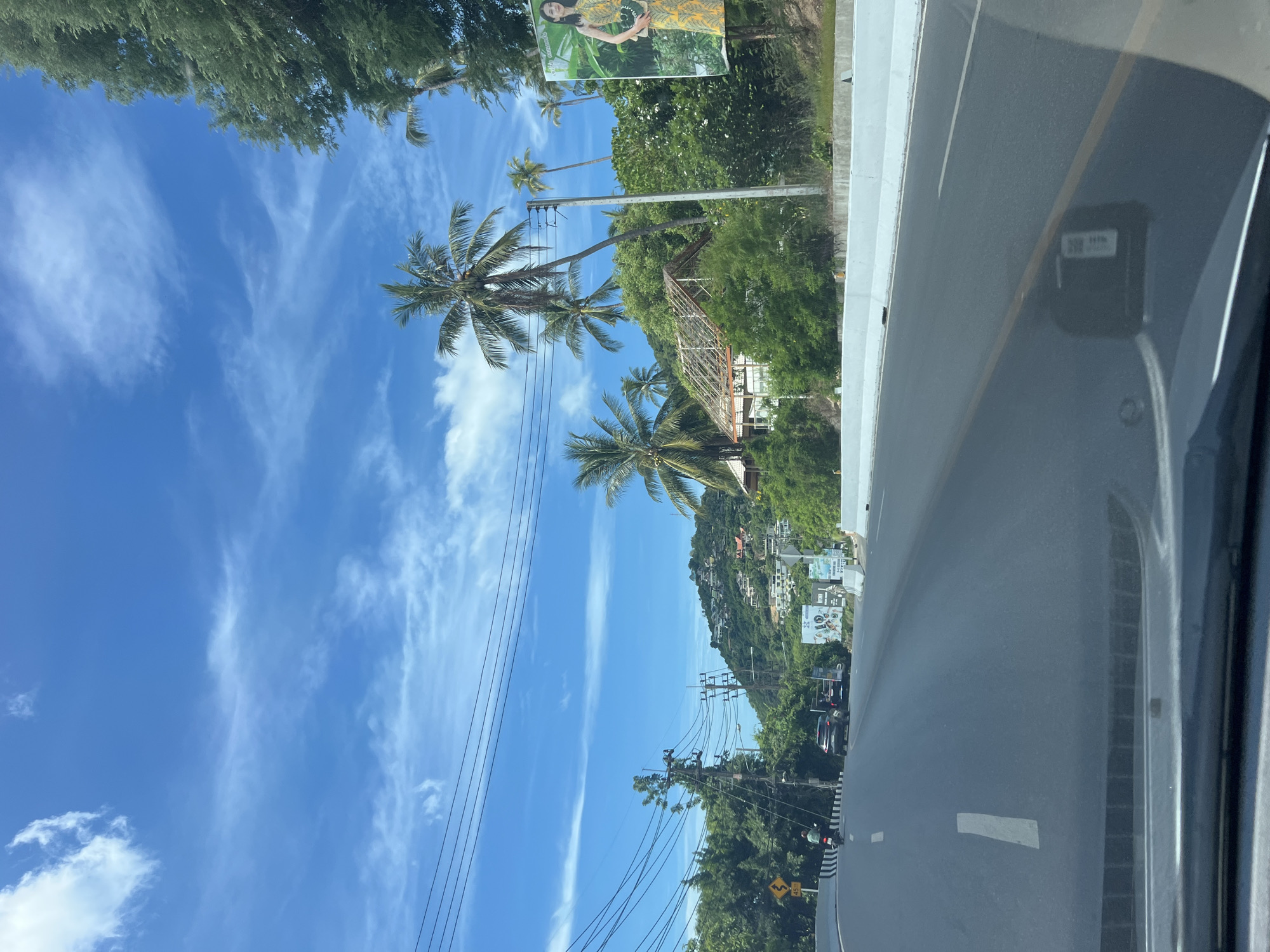 The image depicts a street scene in Baan Chaweng Noi, Thailand, under a bright blue sky with scattered white clouds. The road curves to the left, with a white line marking the lane. A motorcycle is visible in the distance on the left side of the road, along with several cars. Dense green foliage and palm trees line the roadside, with a hillside dotted with houses in the background. A large billboard featuring a woman in a yellow dress is prominent on the right side of the road. Power lines stretch across the sky, supported by utility poles. The scene appears to be captured from the perspective of a moving vehicle.