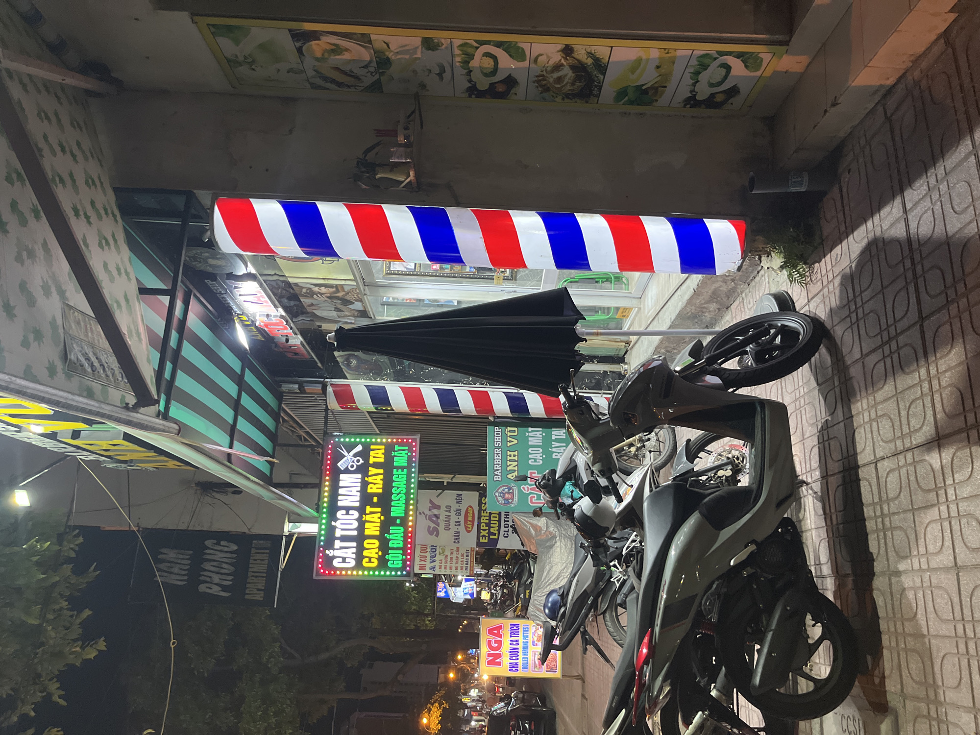 The scene is an outdoor shot in Bac Nha Trang Ward, Vietnam, at night. The focus is on a sidewalk lined with parked motorbikes. Several business signs in Vietnamese are visible. There is a barber pole with red, white, and blue stripes. A brightly lit sign reads "CẮT TÓC NAM CẠO MẶT - RÁY TAI GỘI ĐẦU - MASSAGE MẶT" (which translates to "Haircut Men Shave - Ear Cleaning Wash Head - Face Massage"). Another sign advertises food, reading "NGA CHA CUỐN CẢ TRÍCH (ROLLED HERRING PATTIES)". The environment includes trees and streetlights along the road.