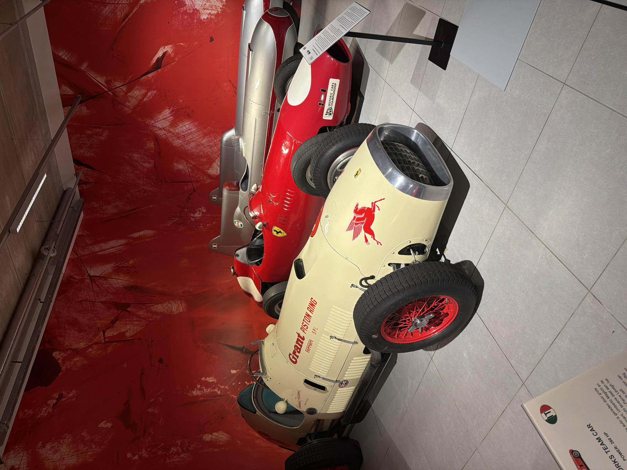 The image shows a museum exhibition of vintage racing cars, with a vibrant red abstract mural as the backdrop. In the foreground, a cream-colored single-seater race car with red detailing on its wheels and body is prominently displayed. The car features the text "Grant PISTON RING" and "FERRARI S.P.L." on its side, along with a winged horse emblem. Behind it, a red Ferrari race car is visible, and further back, two silver streamlined cars are partially seen. A display stand with information is positioned near the red Ferrari. The floor is tiled with large, light grey square tiles. The lighting appears to be artificial, casting subtle shadows. The exhibition is likely located in The Hague, Netherlands, as suggested by context clues and information often found in such museum settings. There are no people visible in the image.