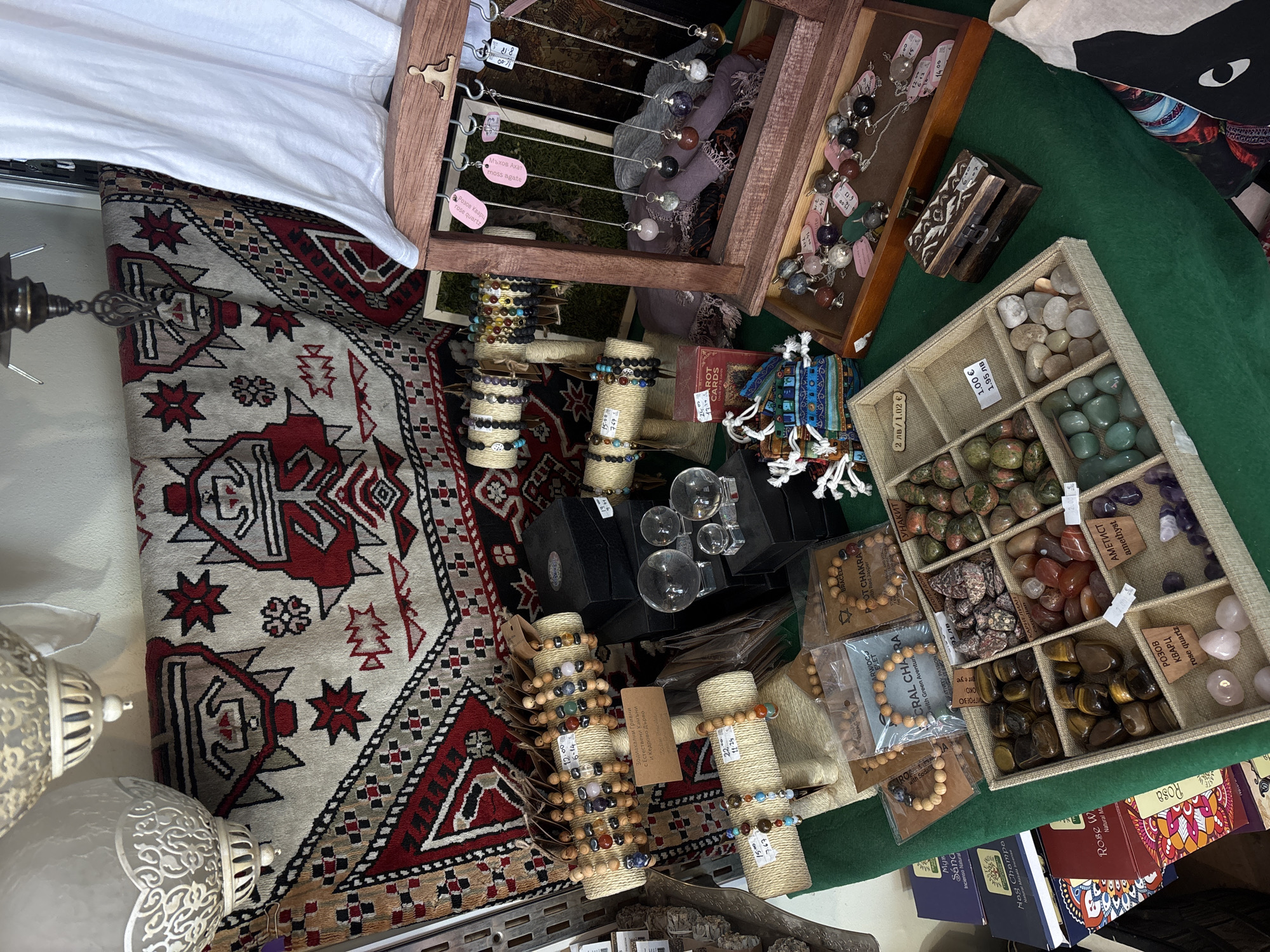 The image depicts a tabletop display of artisan jewelry and related items for sale. The scene includes bracelets made with natural stones and cedar wood, pendulums, and loose gemstones organized in a divided tray. Price tags in both Bulgarian and euros indicate the cost of individual items. There are also stacks of books visible towards the bottom right of the frame. The display is set on a green cloth, accompanied by a patterned carpet, which partially covers the tabletop. The items are densely arranged, showcasing a variety of options for potential customers. The location appears to be a market or fair where handcrafted goods are being sold, possibly in Varna, Bulgaria.