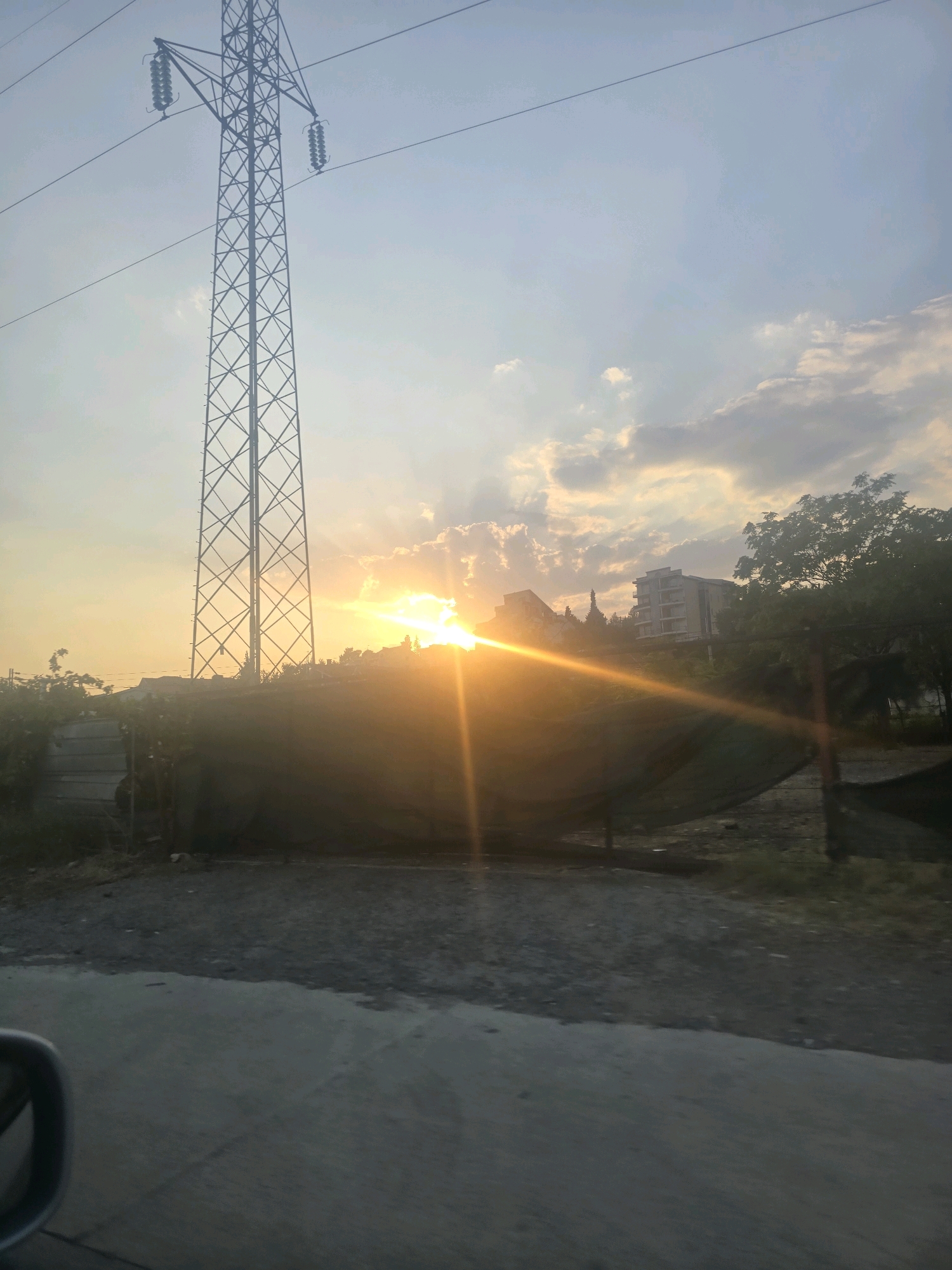 The image depicts a sunset scene with a prominent power line tower in the foreground, situated in Podgorica, Montenegro. The sun is setting behind a cluster of trees and buildings, casting a warm orange glow across the sky and creating lens flares. The tower itself is a tall, lattice-like structure with power lines extending horizontally. Below the tower, there is a large, dark, tarp-like object that covers an area of ground, possibly construction material or a temporary shelter. In the background, several buildings are visible, including what appears to be a multi-story residential building. The overall environment suggests an urban or semi-urban setting with evidence of recent construction or development. The foreground shows a road surface, indicating that the photo was taken from a vehicle.