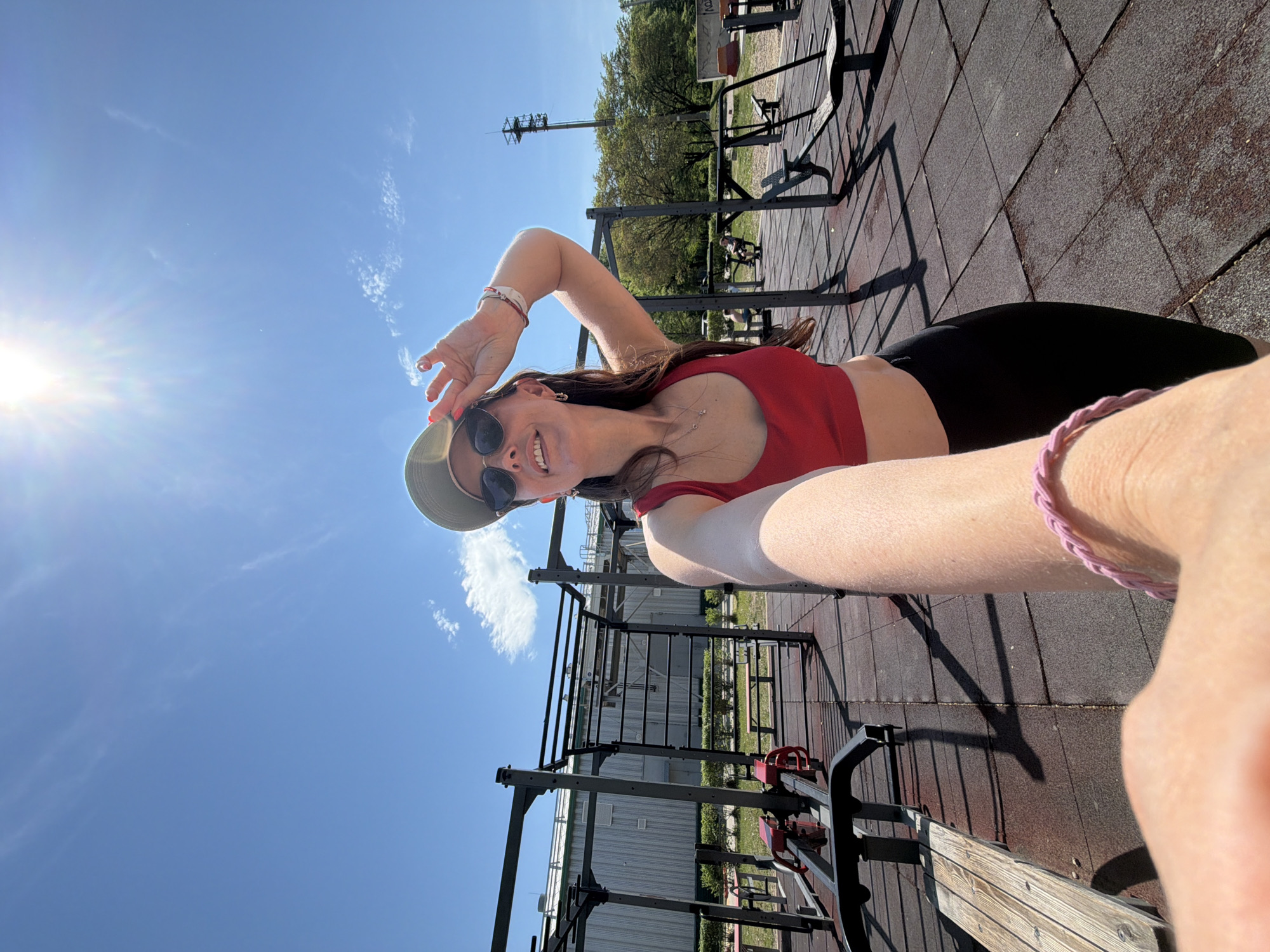 A woman in sunglasses and a hat is taking a selfie outdoors, presumably in Birobidzhan, Russia. She is wearing a red top and black pants. The photo is taken at an angle, showing a blue sky and sunlight. There are outdoor exercise equipment structures in the background, and the ground appears to be made of a rubberized tile material.