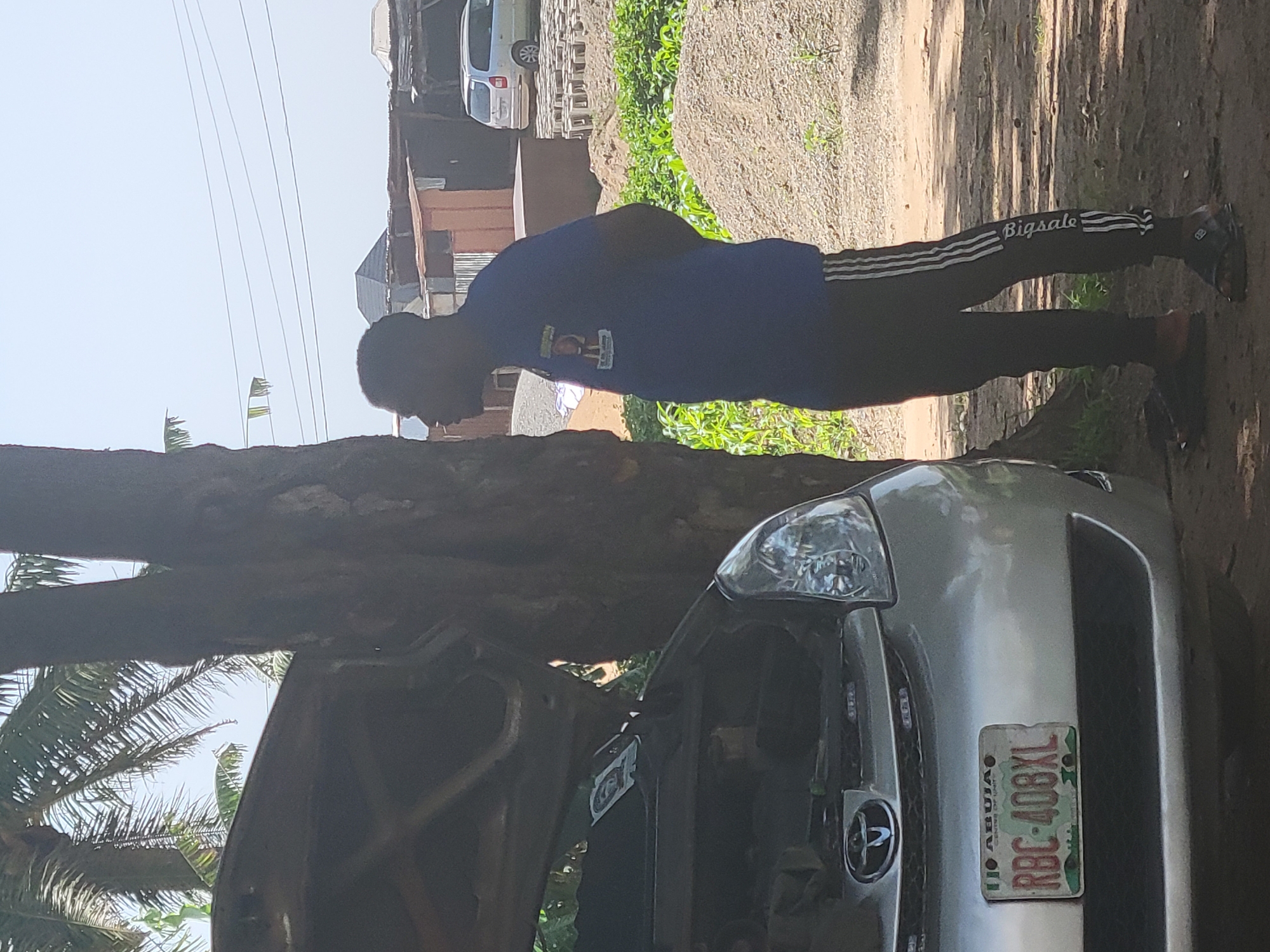 A man in a blue t-shirt and black sweatpants with white stripes stands by an open-hooded silver car. He is facing away from the camera, looking towards the right. The car's license plate reads "RBC-408XL" and displays "ABUJA CENTRE OF UNITY". Behind the man and car is a dirt area with piles of gravel, some green plants, and scattered concrete blocks. In the background, there are simple structures made of corrugated metal and wood, as well as a light-colored minivan. Power lines stretch across the sky. A palm tree trunk is visible to the left of the man and the car. The scene appears to be outdoors, possibly in an informal auto repair or storage area, in Suleja, Nigeria.