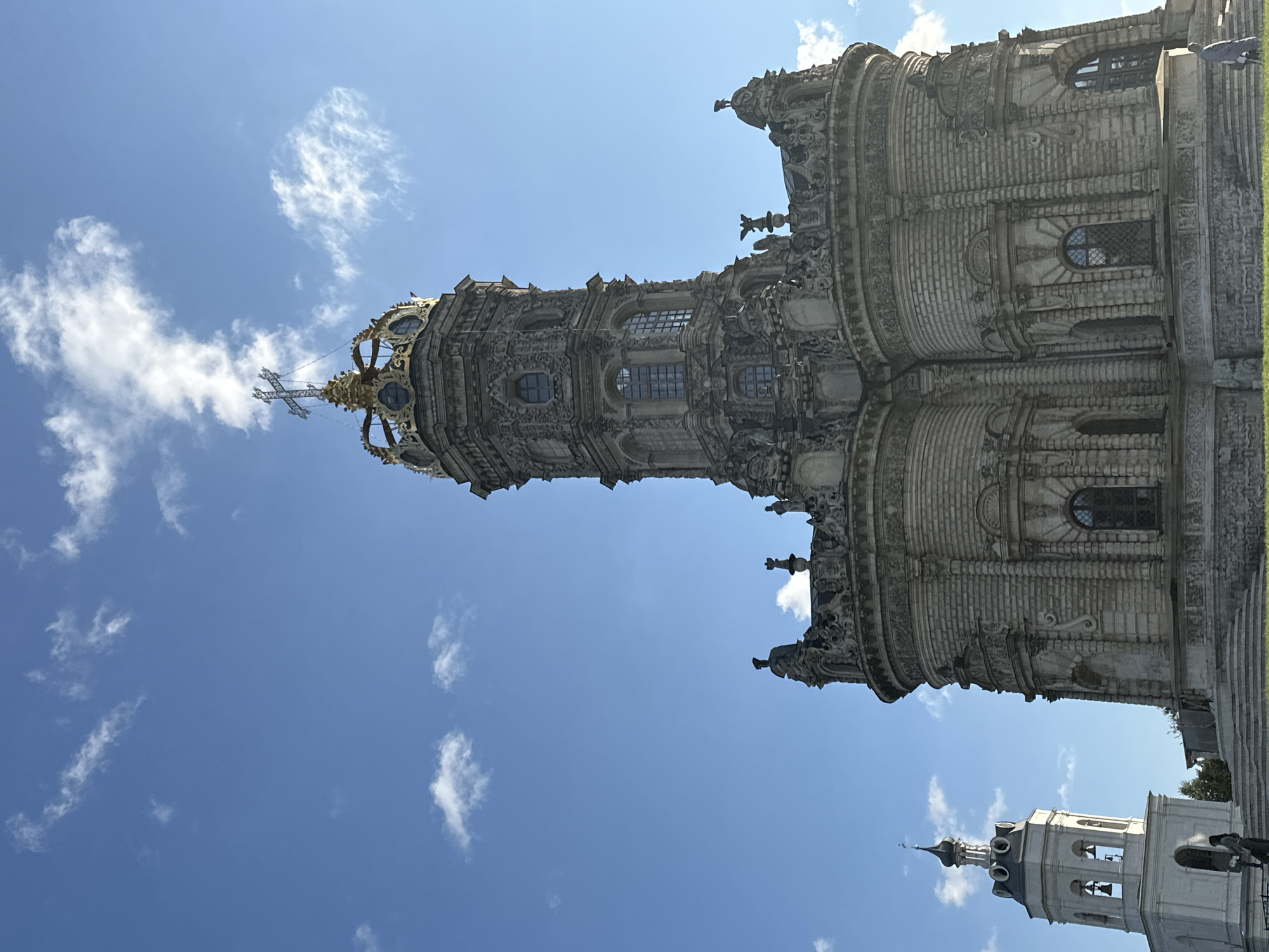 The image shows the Church of the Sign of the Theotokos in Dubrovitsy, Russia, under a clear blue sky with scattered white clouds. The church is a Baroque style building with intricate stone carvings and a distinctive golden crown-like dome topped with a cross. To the left of the main church, a white bell tower with a dark spire is visible. A person is walking up a set of stairs in front of the church. The grounds around the church appear to be green and grassy.