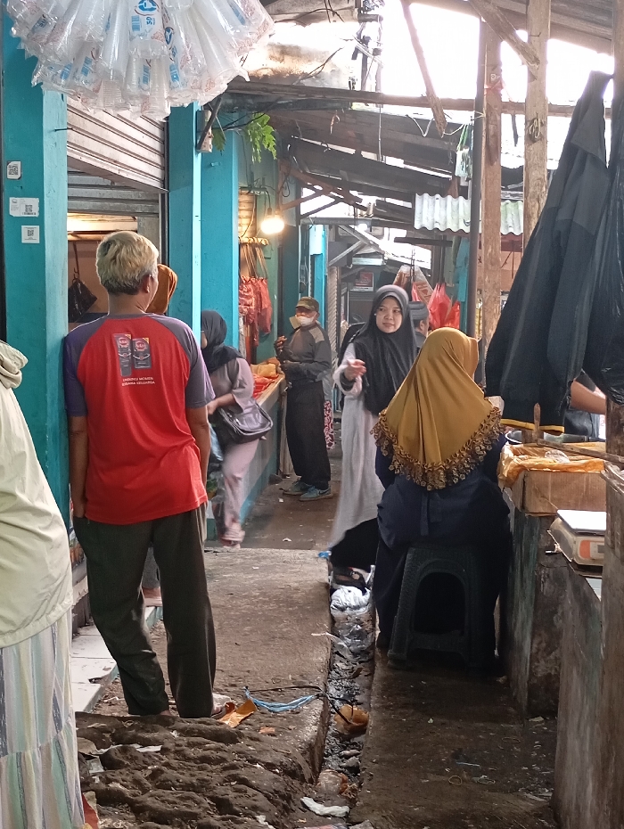 The image shows a narrow street in Kutawaringin, Indonesia, where people are walking, shopping, and talking. The location is a narrow alleyway, with wooden structures and concrete walls. The alleyway is cluttered with loose items. There are six people in the image, all wearing typical Indonesian clothing. One person is wearing a red shirt and appears to be talking to someone. Another person is wearing a yellow headscarf, sitting on a stool, and looking to the right. A third person is wearing a blue headscarf and holding a bag. A fourth person is wearing a black headscarf and is looking at the camera. A fifth person is wearing a white headscarf and is walking past. A sixth person is wearing a blue shirt and is walking toward the camera. The image was captured in a brightly lit environment, which seems to be during the day. There is a dark, black umbrella on the right side of the image, and a bag on the floor to the left of the image. The alleyway is dirty, with dirt and debris scattered on the ground. The scene is a casual and informal setting, and the people appear to be engaged in everyday activities.