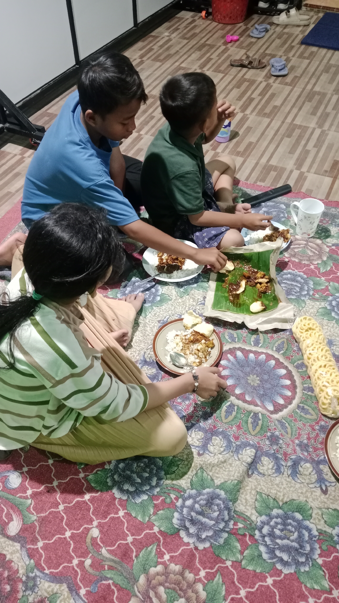 The image depicts a family meal scene, likely taking place indoors on a patterned rug. Three individuals, appearing to be two boys and a girl, are seated on the floor, engaged in eating. The boys are positioned on the left and center, while the girl is on the far right. They are surrounded by plates of food, rice, and what appear to be crackers, some served on banana leaves. A white mug is also visible on the right. The environment suggests a casual home setting, possibly in the city of Madiun, Indonesia, given the cultural context often associated with such meal arrangements. The individuals are dressed in casual attire, with the girl wearing a striped long-sleeved shirt and the boys in t-shirts. The focus is on the shared meal and the familial interaction.