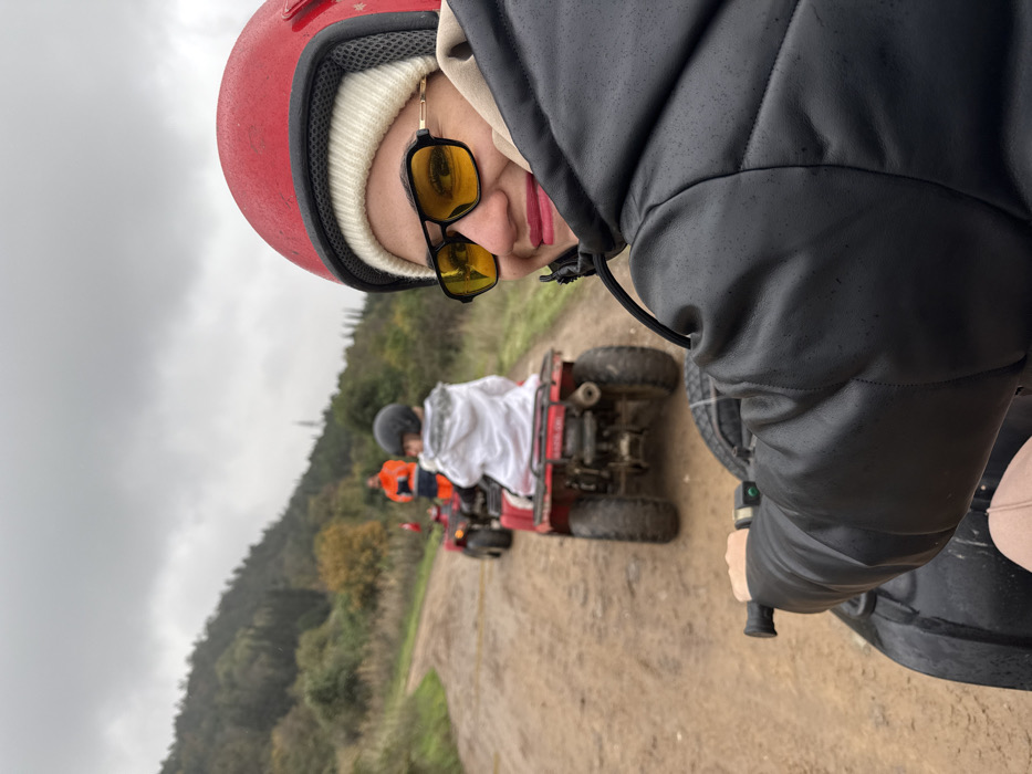 The image depicts a group of three individuals riding ATVs on a muddy dirt path in a rural, hilly area within Çiftlikköy, Turkey, under an overcast sky. In the immediate foreground, the right side of an individual is visible, wearing a red helmet over a white beanie, black-framed sunglasses with yellow lenses, a black padded jacket, and holding the black handlebar of an ATV. This person has pink lipstick and is smiling, looking towards the viewer. Directly in front, a second person, appearing to be a child, rides a red, mud-splattered ATV, wearing a black helmet and a white patterned jacket. Further along the winding dirt path, a third person on another ATV is visible, wearing an orange jacket or vest. The environment consists of green, tree-covered hills in the background and sparse vegetation flanking the unpaved path.
