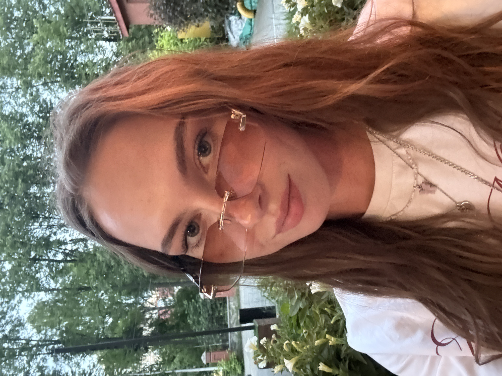 A young adult female is centered in a close-up, head-and-shoulders portrait, appearing to be a selfie taken outdoors. She has long, wavy, light brown hair with reddish undertones, partially obscuring her shoulders. Her light-colored eyes (appearing blue or gray) are visible above rimless sunglasses with wide, rectangular, light brown tinted lenses, framed by slender gold-toned hardware at the temples and bridge. She displays a slight, closed-mouth smile. She wears a white long-sleeved top or sweatshirt, with a faint maroon design visible on the left chest/shoulder area. Multiple silver-colored chain necklaces are visible; one features a small, light pink, butterfly-shaped charm, and another has a small, round, silver-toned pendant.
The background is an outdoor, verdant setting. Lush green trees with dense foliage are prominent in the upper and mid-ground, suggesting a park or garden environment. To the left, a dark vertical pole or post is visible. On the right, part of a building with a reddish-brown roof and light-colored walls is discernible, partially obscured by dark green bushes. In the foreground, particularly on the left, white petunia-like flowers and green leafy plants are in bloom. The lighting is soft and diffused, indicating overcast conditions or late day light. No specific visual cues, such as landmarks or discernible text, identify the location as Perm, Russia.
