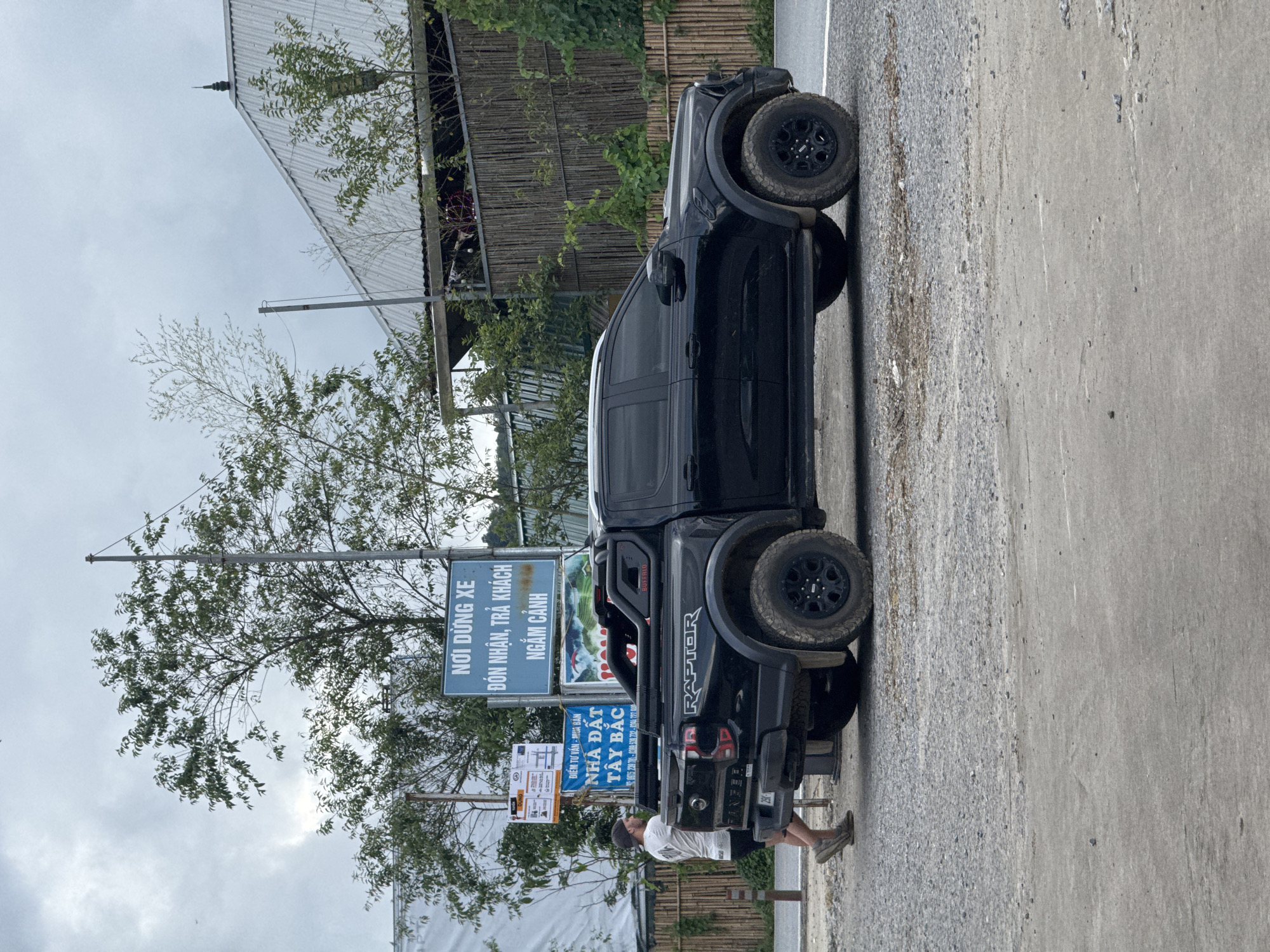 This image captures a man standing beside a black pickup truck in Tu Le, Van Chan District, Yen Bai Province, Vietnam, specifically near a location designated for parking, passenger drop-off/pick-up, and sightseeing, as indicated by the blue sign reading "NƠI DỪNG XE ĐÓN NHẬN, TRẢ KHÁCH NGẮM CẢNH."
The man, with short dark hair and a baseball cap, wears a white t-shirt, dark shorts, and athletic shoes. He is positioned on the left side of the truck's rear, facing away from the camera, appearing to interact with or stand near the vehicle.
The vehicle is a black Ford Ranger Raptor pickup truck, identifiable by the "RAPTOR" branding on the bed and "RANGER" on the tailgate. It features a black roll bar or sport bar in the truck bed with "BUFFALO" written on it. The truck is equipped with aggressive off-road tires and black rims, and its body appears somewhat dirty, suggesting recent travel on unpaved surfaces. The rear license plate is partially obscured but shows "29K".
Behind the man and the truck, two informational signs are visible. The prominent blue sign is for the parking and passenger area. Below it, a smaller sign advertises "NHÀ ĐẤT TÂY BẮC" (Northwest Real Estate) with Vietnamese phone numbers. Further to the left, a white sign with various smaller labels, possibly directions or regulations, is partially visible.
The ground surface in the foreground and beneath the truck is a mix of gravel and packed dirt/concrete. In the background, there are numerous trees with green foliage and utility poles. A structure with a corrugated metal roof and a bamboo-slat fence is visible behind the trees and the truck, suggesting a building or a viewing platform. The sky is overcast and grey, indicating a cloudy day.