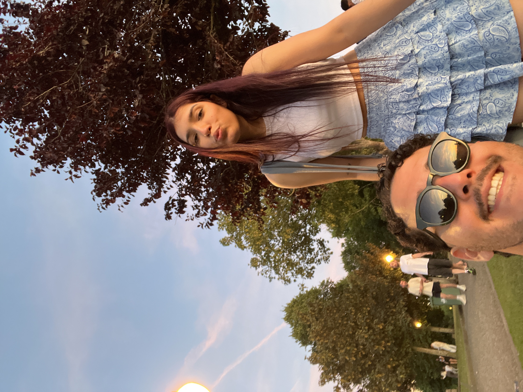 This is a selfie taken outdoors, likely in a park in Vila Nova de Gaia, Portugal, as suggested by the presence of trees, pathways, and people engaged in leisure activities. The sky is a gradient of pale blue with wispy pink clouds, indicative of either dawn or dusk. A large, dark-leafed tree dominates the upper right portion of the frame.
In the foreground, the lower half of a man's face is visible, wearing sunglasses that reflect the sunset sky. He has curly dark hair and a mustache, and he is smiling. Behind him and slightly to the left, a woman is positioned. She has long, dark hair with purple undertones and is making a kissing face. She is wearing a white crop top and a blue, patterned ruffled skirt. She also has a grey bag slung over her shoulder.
In the background, several people are visible walking along a pathway. Two men are walking side-by-side, one in a light-colored t-shirt and shorts, the other in a white t-shirt and dark shorts. Other figures are more distant and less distinct. Several lampposts are scattered throughout the background, one of which is prominently lit on the left side of the image. The ground appears to be a mix of grass and paved paths.