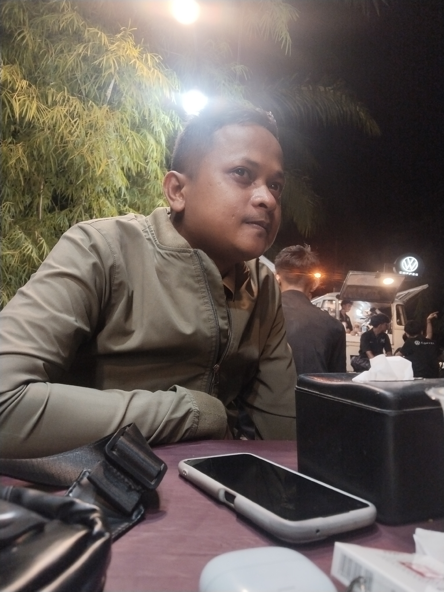 The image depicts a man sitting at a table outdoors at night, likely at a roadside cafe or food stall. He is wearing a khaki bomber jacket. On the table in front of him are a smartphone, a tissue box, and a bag with a buckle. In the background, a food truck with a "COFFEE" sign is visible, along with other people. The setting appears to be a casual, urban environment, consistent with a location in Lhokseumawe, Indonesia.