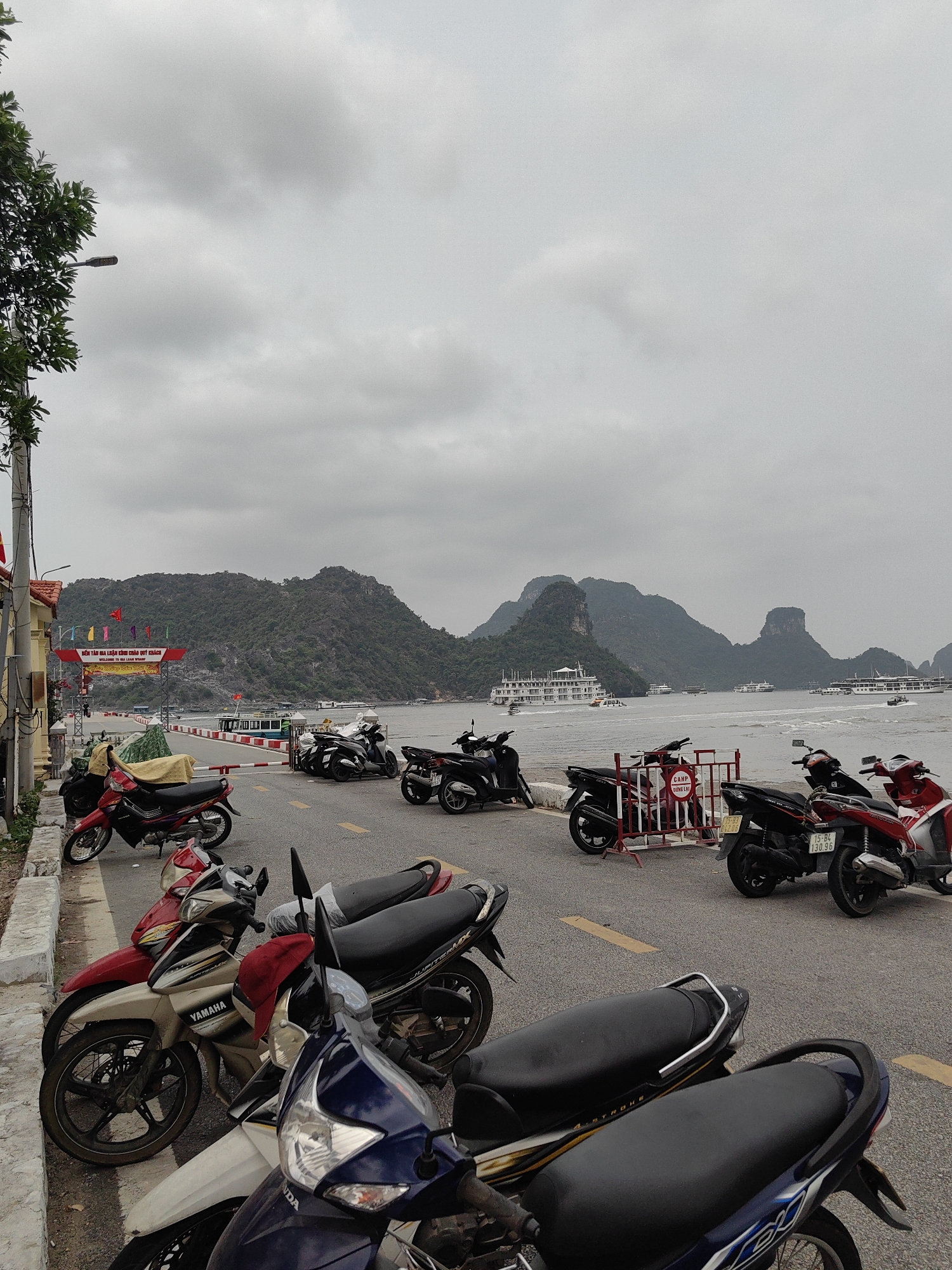 The image depicts a street scene in Hải Phòng, Vietnam, filled with parked motorcycles on a paved road marked with yellow lines. Beyond the motorcycles, there is a body of water with several islands or karst formations visible in the distance. A few large cruise ships are anchored or navigating on the water. The sky is overcast with gray clouds. On the left side of the image, there is a sidewalk with a building and some vegetation. A sign in Vietnamese is visible above the road. No people are distinctly discernible in the image, although some may be obscured by the motorcycles or distant in the scene.