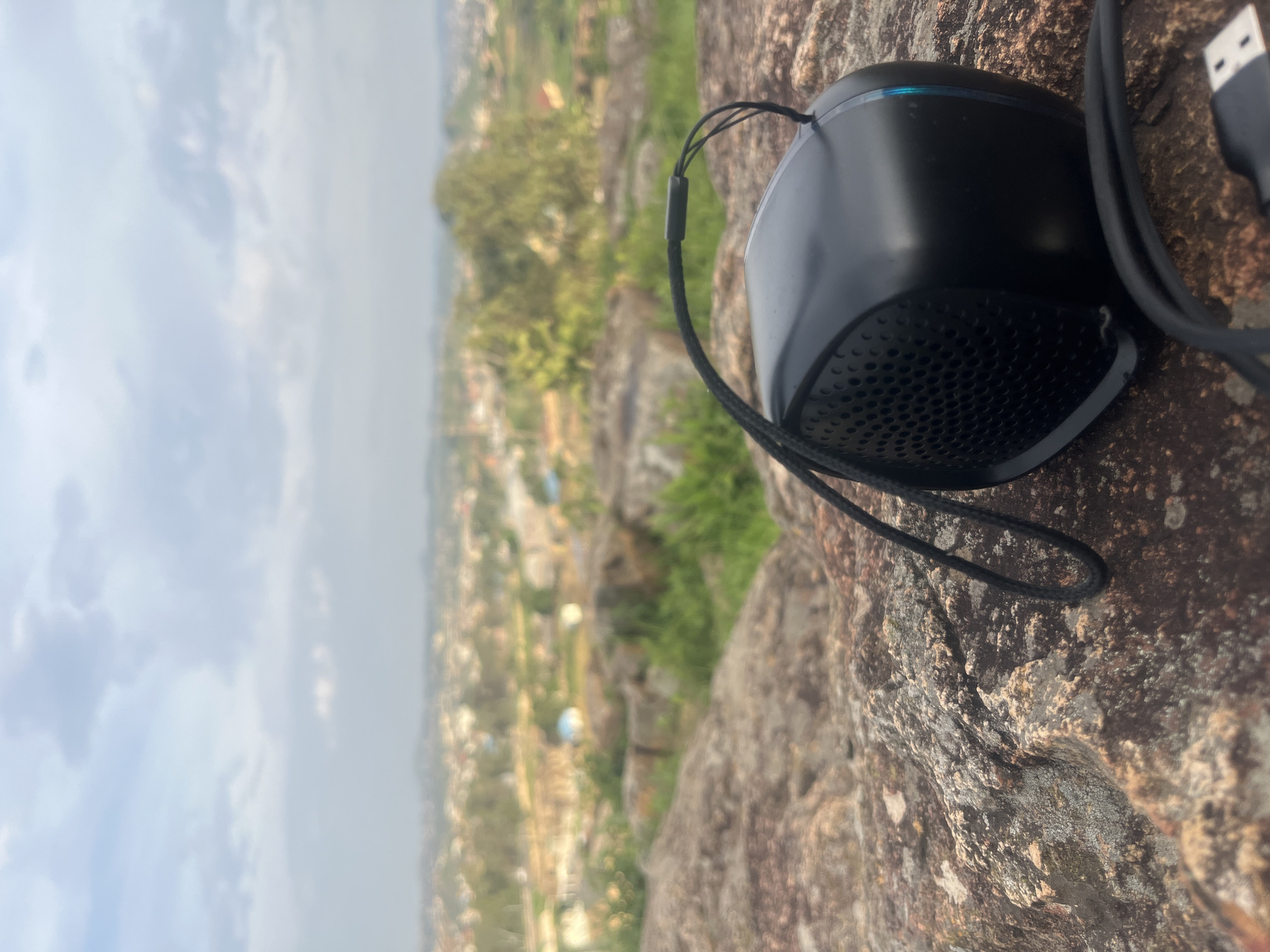 A black portable speaker with a black cord and a strap is positioned on a rocky surface in the foreground, overlooking a city in Gura Top, Nigeria. The speaker is facing the viewer's right, with its perforated grille clearly visible. A USB cable is also present, extending from the speaker. The background shows a hazy cityscape with buildings and greenery under a cloudy sky. The rocks in the foreground are textured and appear to be part of a natural landscape. The lighting suggests it is daytime.