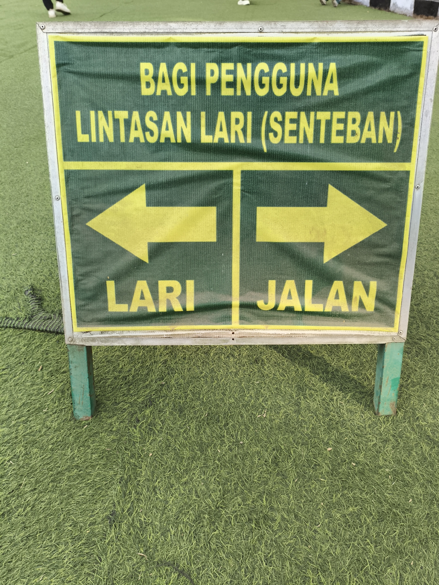 The image shows a green and yellow sign on a grassy field, likely artificial turf, with metal posts supporting it. The sign is divided into two sections by a vertical yellow line. The top part of the sign has text in yellow: "BAGI PENGGUNA" above "LINTASAN LARI (SENTEBAN)". The bottom left section has a yellow arrow pointing left and the word "LARI" in yellow. The bottom right section has a yellow arrow pointing right and the word "JALAN" in yellow. The sign is framed by a metallic border. In the background, parts of people wearing shoes are visible, suggesting they are on the field. A black rope is visible on the left side of the sign, extending onto the grass. The scene appears to be an outdoor sports or recreational area. This sign indicates designated paths for running and walking. This scene is from Magelang, Indonesia.