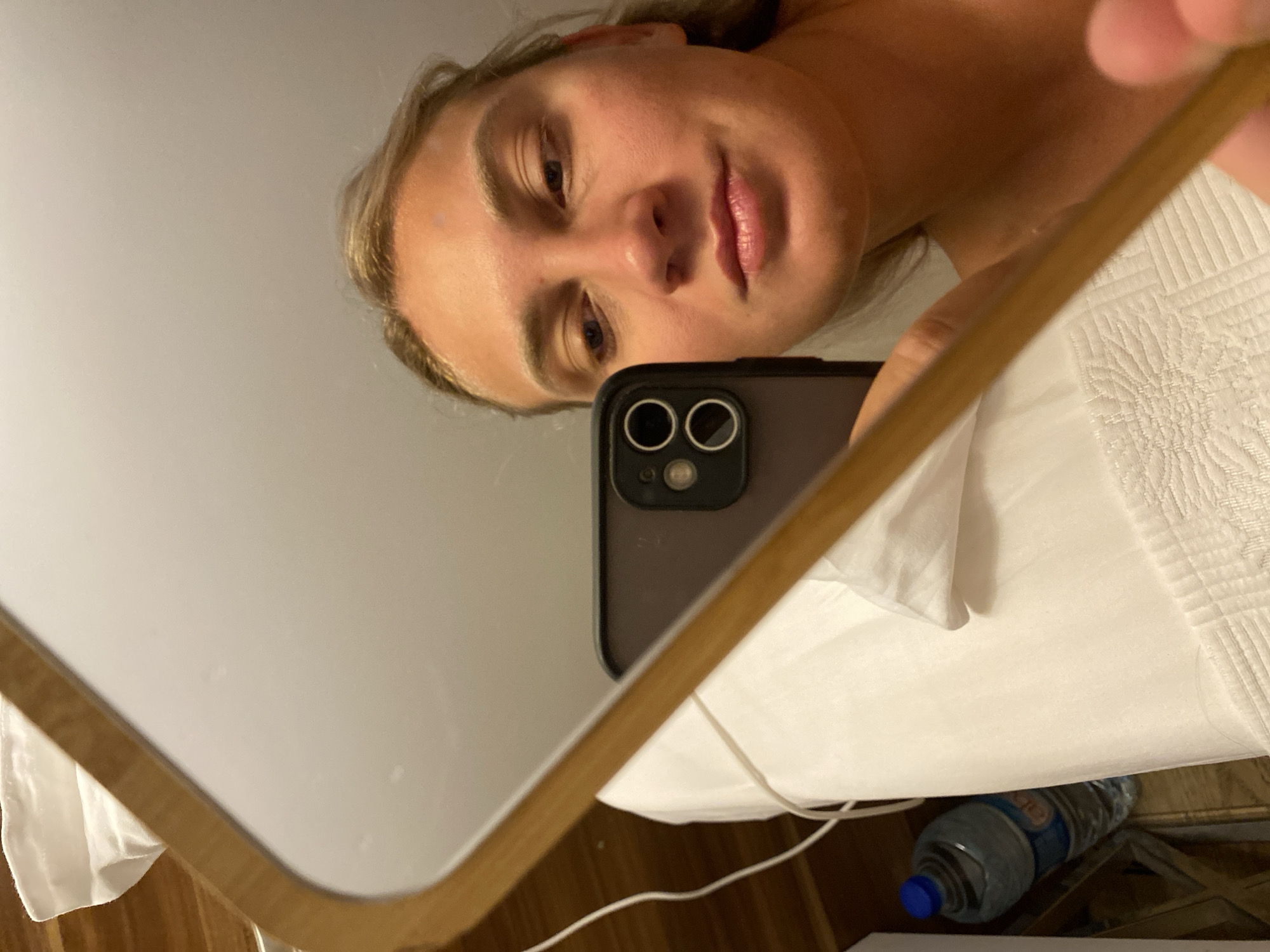 A close-up, low-angle, mirror selfie is captured from the perspective of a person holding a smartphone. The person's face, partially obscured by the phone, is visible in the reflection. They have light-colored hair and fair skin. The background of the reflection shows a white surface, possibly a bed or towel, with a bottle of water in the lower left corner. The mirror itself has a wooden frame and is angled downwards. The surrounding environment appears to be indoors. The image's metadata does not contain location information.