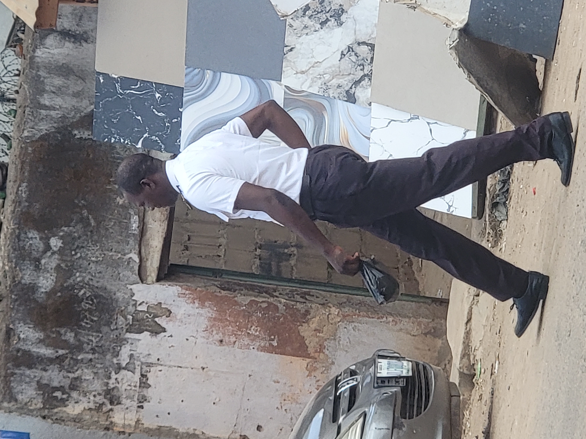 An adult male dressed in a white short-sleeved shirt and dark trousers is seen in the city of Maiburiji, Nigeria. He is bent over, holding a dark object in his left hand. The backdrop features a weathered building with exposed brick and peeling paint, alongside a display of tile samples. A portion of a silver vehicle is visible in the foreground. The scene is located outdoors on a surface that appears to be a street or sidewalk.