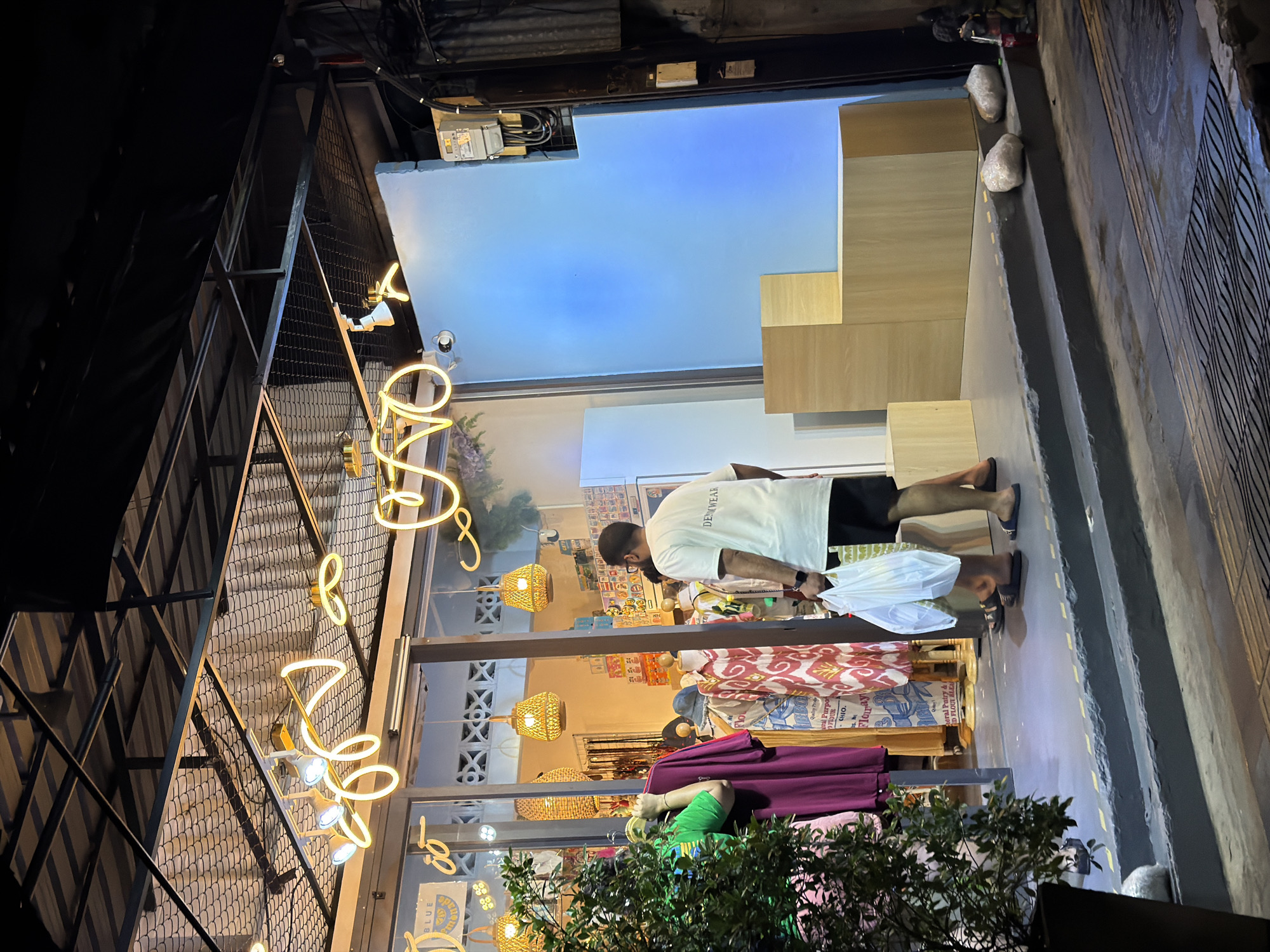 A man in a white t-shirt and black shorts is standing outside a shop, holding two plastic bags. The shop appears to be a clothing store, with garments displayed in the window. Wicker pendant lights hang inside the store. Neon signs are visible on the exterior of the building. The scene is illuminated by artificial lighting, suggesting it is nighttime. The man is facing away from the camera, towards the shop entrance. The background shows a blue wall and wooden shelving units. The setting appears to be a street in Phuket City Municipality, Thailand.