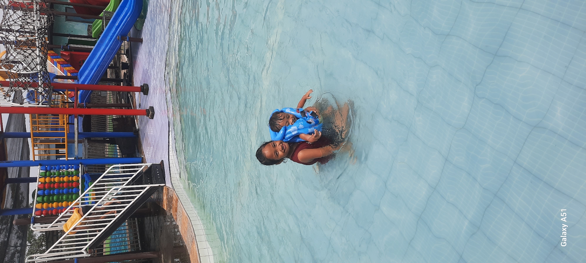 In the city of Popoh, Indonesia, two young females are in a swimming pool. The older female, wearing a dark red sleeveless shirt, holds the younger female in her arms. The younger female is wearing a blue inflatable life vest. The water in the pool is clear and shallow, with a tiled bottom. In the background, there is a colorful water park structure with slides and climbing equipment. The overall scene depicts a fun and safe aquatic environment.