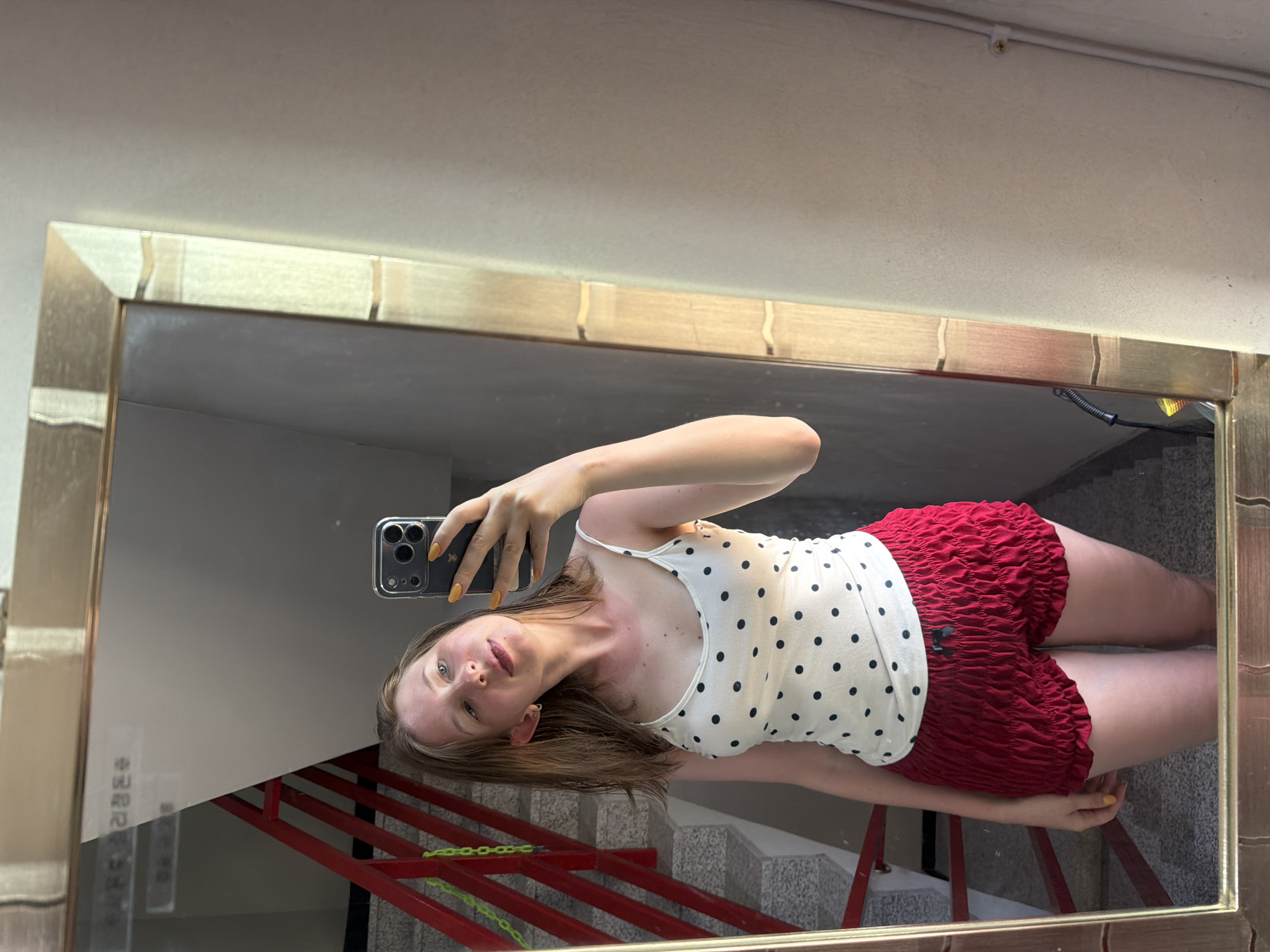 A young woman with shoulder-length brown hair takes a mirror selfie. She is wearing a white tank top with black polka dots and red ruffled shorts. She is holding a smartphone in her right hand and looking at the camera. The mirror reflects a staircase with red railings and a grey wall. The scene appears to be indoors, possibly in a residential building. The text on the wall visible in the mirror is in Korean. Based on the context of the Korean text, this location is likely in Gangneung-si, South Korea.