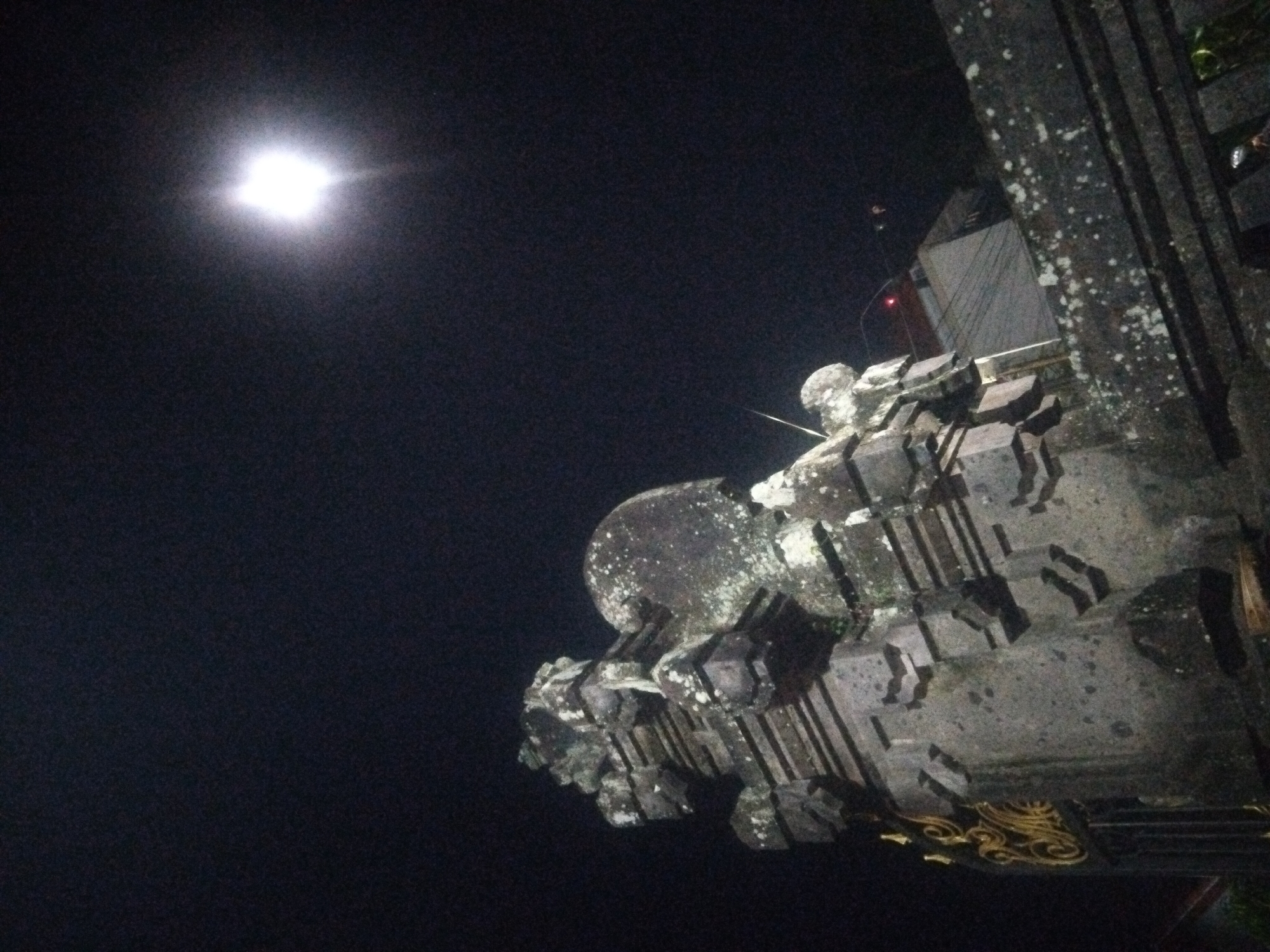 Captured from a low angle at night in Unknown, Unknown, the photograph features a stone structure in the foreground. The structure is ornamented with intricate carvings and displays signs of weathering, as evidenced by the presence of white lichen or mold. A bright, circular light source, likely a streetlamp or the moon, dominates the dark sky in the upper left corner of the frame. To the right of the stone structure, there appears to be a building or a wall, partially illuminated, and vertical wooden beams. The overall scene conveys a sense of stillness and mystery due to the darkness and the historical appearance of the stone structure.