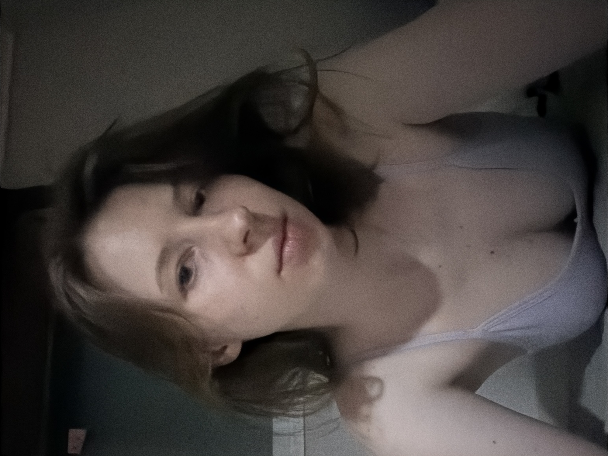 The image is a close-up, low-angle selfie of a young woman with light brown hair and fair skin. She is looking towards the camera with a neutral expression. She is wearing a light-colored tank top or bra. The background appears to be an indoor setting with muted lighting, and a dark wall or surface behind her. The image is grainy and has a slightly blurred quality. The exact location within Dongducheon-si, South Korea, is not determinable from the visual information provided in the image.