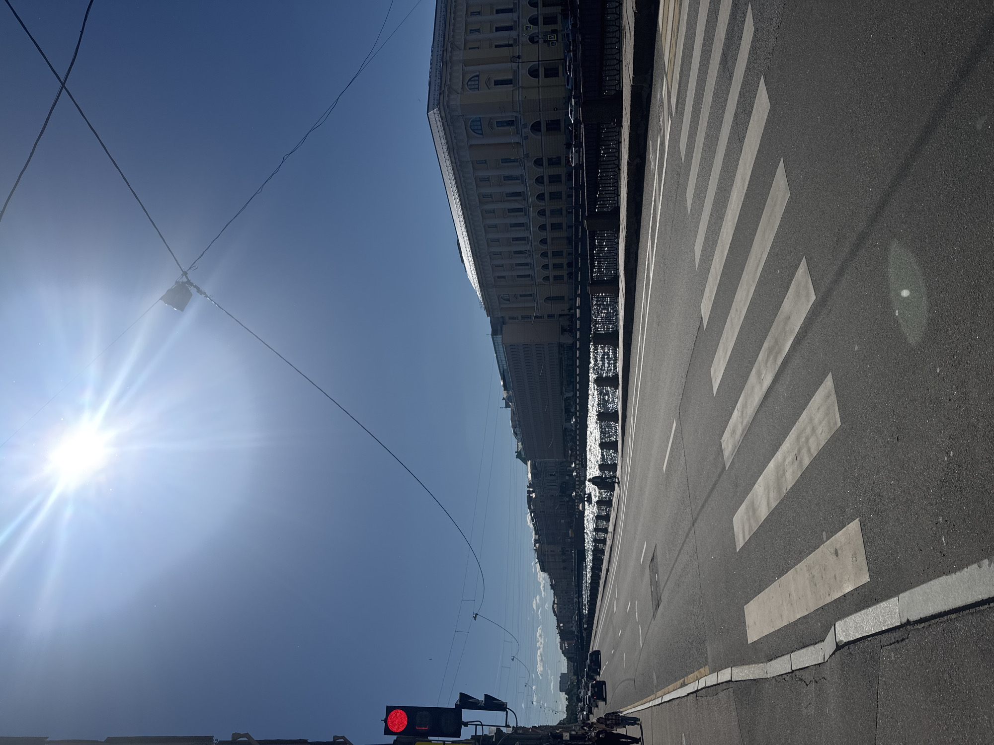 The image depicts a sunny day in Saint Petersburg, Russia, likely along a wide boulevard adjacent to a canal or river. The viewpoint is from the street, looking towards the opposite embankment.
In the foreground, a pedestrian crosswalk is clearly visible, marked with wide white stripes on dark asphalt. To the left, the sidewalk is bordered by a curb and the street continues, with a traffic light showing a red signal. Several cars are parked and moving along the street. Two pedestrians are walking on the sidewalk to the left.
The middle ground is dominated by a shimmering body of water, reflecting the bright sunlight. Along the embankment of the water, a decorative metal fence runs, separating it from a row of multi-story buildings. These buildings exhibit classical architectural styles, with large windows and columns.
The background features a clear blue sky with the sun prominently positioned in the upper left quadrant, casting strong rays and a lens flare. Overhead, power lines and cables are strung across the sky. The overall scene is one of a bustling urban environment on a bright, clear day.
