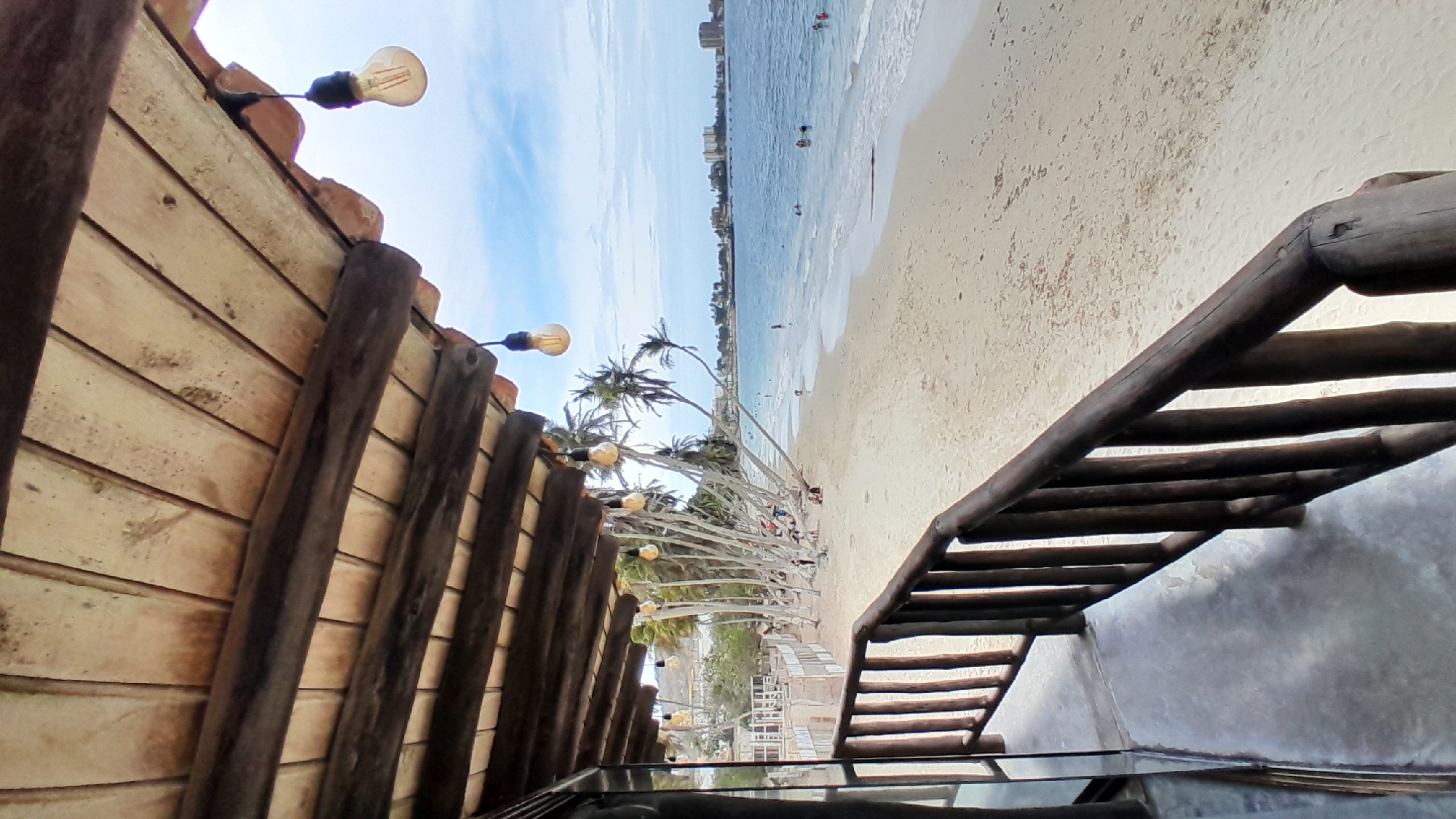 The image presents a detailed view from under a wooden veranda or balcony overlooking a sandy beach and the Caribbean Sea in Pampatar, Venezuela.
In the foreground, the structure from which the photo is taken features a rustic wooden ceiling composed of light-colored planks supported by darker, thicker wooden beams. Several vintage-style incandescent light bulbs, suspended by dark cords, hang from this ceiling. The edge of a tiled roof, with reddish-brown undulations, is visible along the top right perimeter. A dark, reflective vertical surface, possibly a window frame, is partially visible on the far left. A dark wooden railing with vertical slats and a horizontal handrail runs diagonally across the lower right portion of the image, suggesting an elevated viewpoint. The floor beneath the railing is a light gray concrete surface.
The midground reveals a wide expanse of light beige sand, forming a curved beach shoreline. Numerous slender palm trees, many leaning distinctly towards the ocean, line the back edge of the beach, providing scattered shade. Several individuals are present on the beach; some are seated or lounging near the base of the palm trees, while others are closer to the water's edge. Further down the beach to the left, dilapidated or rustic structures are visible.
The background features the calm, blue waters of the ocean, with gentle white foamy waves breaking near the shore. Multiple people are scattered in the water, swimming or wading. In the distant background, across the bay, a low-rise urban landscape with various buildings and structures is visible under a slightly hazy sky. The sky above is a light blue with scattered white, fluffy clouds. The overall lighting suggests a bright daytime scene.