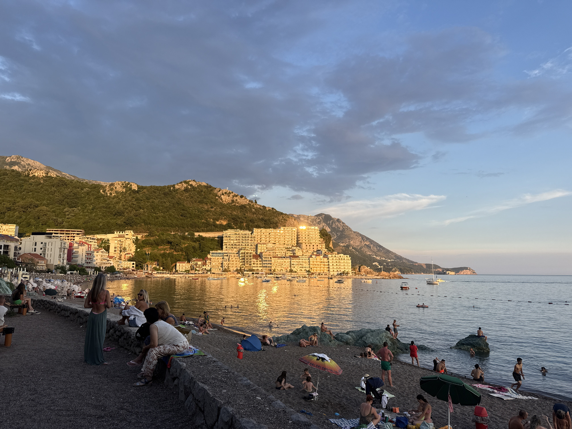 This media file depicts a lively beach scene at Rafailovići, Montenegro, during what appears to be late afternoon or early evening. The golden hour sunlight illuminates the buildings lining the coast and casts a warm glow on the calm sea.
The foreground is occupied by a dark-pebble beach where numerous people are enjoying various activities. Some are relaxing on towels, others are sitting on low stone walls, and a few are wading in the shallow water. There are several beach umbrellas, a few small inflatable toys, and beach bags scattered across the sand. A woman in a long, flowing green dress stands out on the walkway, facing away from the camera.
Further back, the sea is dotted with several small boats and a sailboat, suggesting leisurely maritime activity. A chain of buoys marks a designated swimming area. The background features a dense cluster of multi-story buildings that ascend the green, mountainous terrain, characteristic of coastal Mediterranean towns. The sky is a mix of blue and dramatic, dark clouds, hinting at the possibility of changing weather. The overall mood is one of relaxed enjoyment and vacation. No visible text is present in the image.
