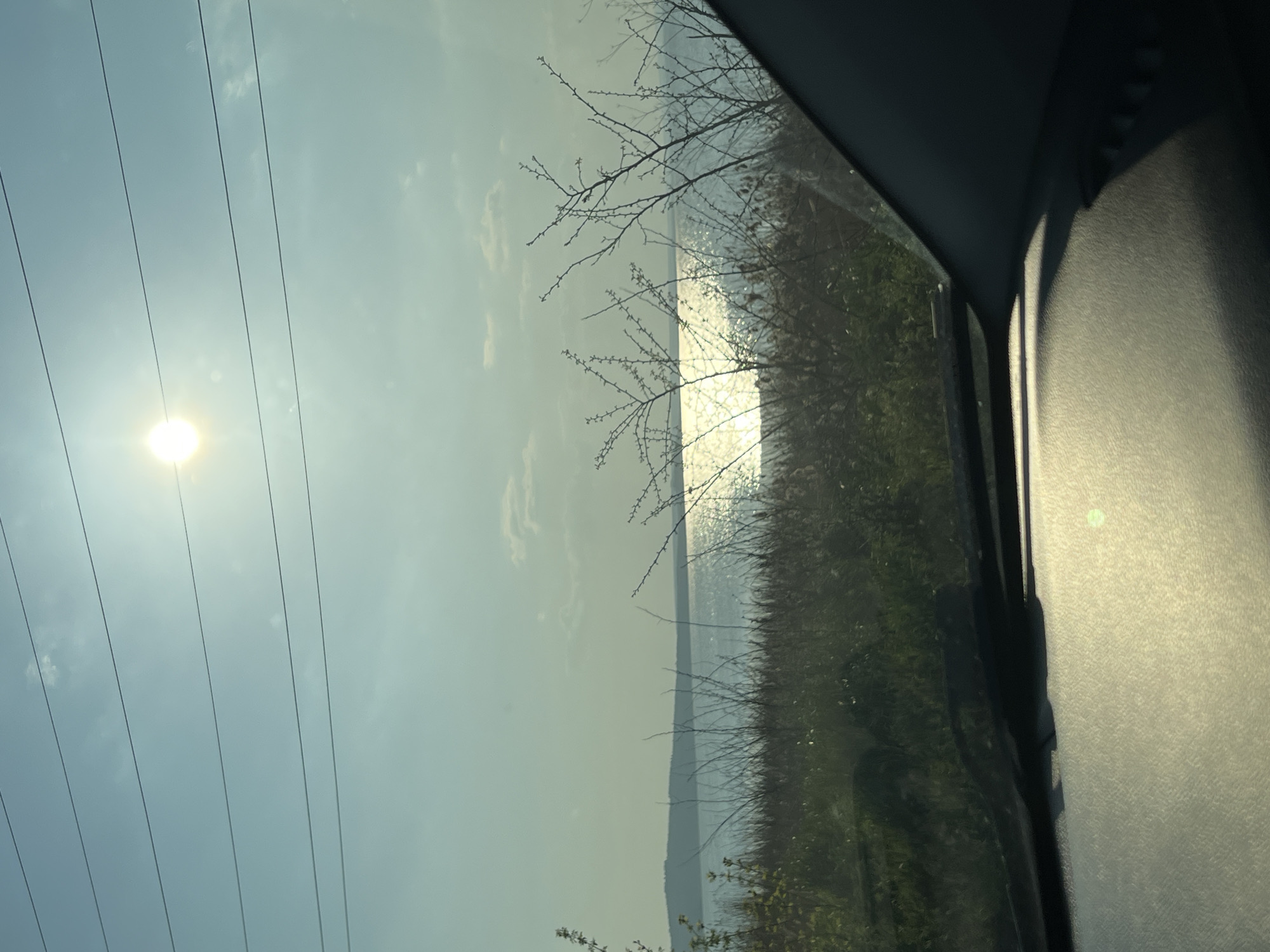The image shows a view from inside a vehicle, possibly a car. On the left, there is a view of the sky with power lines and the sun shining brightly. In the center, there is a body of water with shimmering reflections of the sun. Some vegetation or grass can be seen along the water's edge, along with bare branches. The right side of the image shows the interior of the car. There are no people visible. The location appears to be near a body of water, potentially a lake, river, or the sea. It is not possible to determine the location as Burgas, Bulgaria from the image alone.