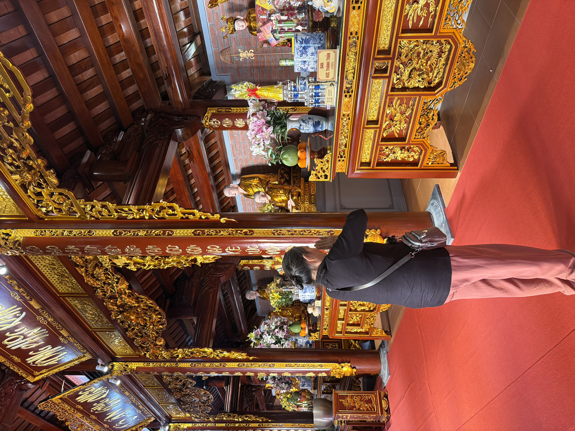 A woman with dark hair is praying in what appears to be a Buddhist temple. She is wearing a black shirt and rose-colored pants. A small purse is visible across her shoulder. The temple's interior features red carpets and elaborately decorated wooden structures, with gold accents. In the background, there are statues and offering tables with flowers and other religious items. The temple has columns covered in gold lettering. The location is Diên Khánh, Vietnam.