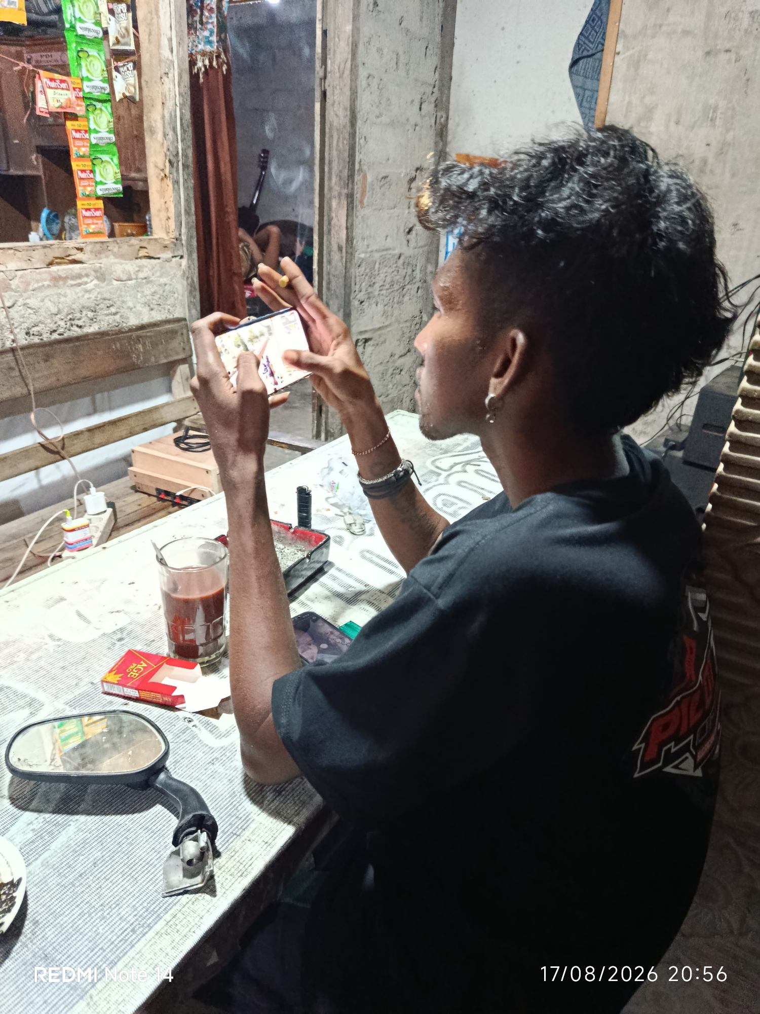 The image depicts a young man with dark, curly hair and an earring, sitting at a table and playing a mobile game. He is wearing a black t-shirt with a red and white graphic on the back. On the table in front of him, there is a glass of dark liquid, a pack of cigarettes, an ashtray with cigarette butts, and a motorcycle mirror. In the background, there's a window with various packets of instant beverages hanging from it. The overall setting appears to be a humble interior, possibly a home or a small shop, in Panggungrejo, Indonesia, as indicated by the date stamp "17/08/2026 20:56" and the watermark "REDMI Note 14". The lighting suggests it is nighttime or a dimly lit indoor space.